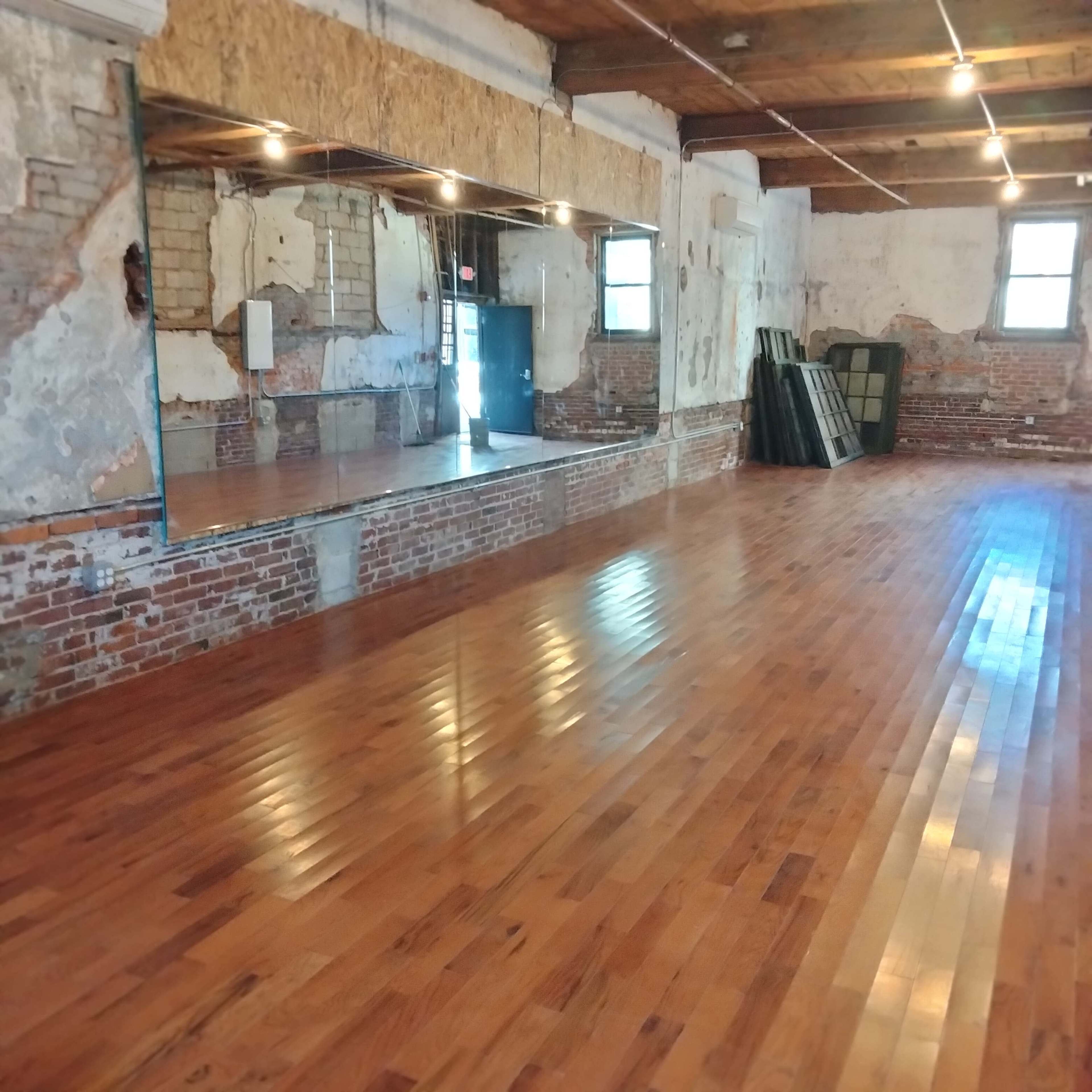 The image shows an empty studio space with polished wooden floors, exposed brick walls, and large mirrors on one side.