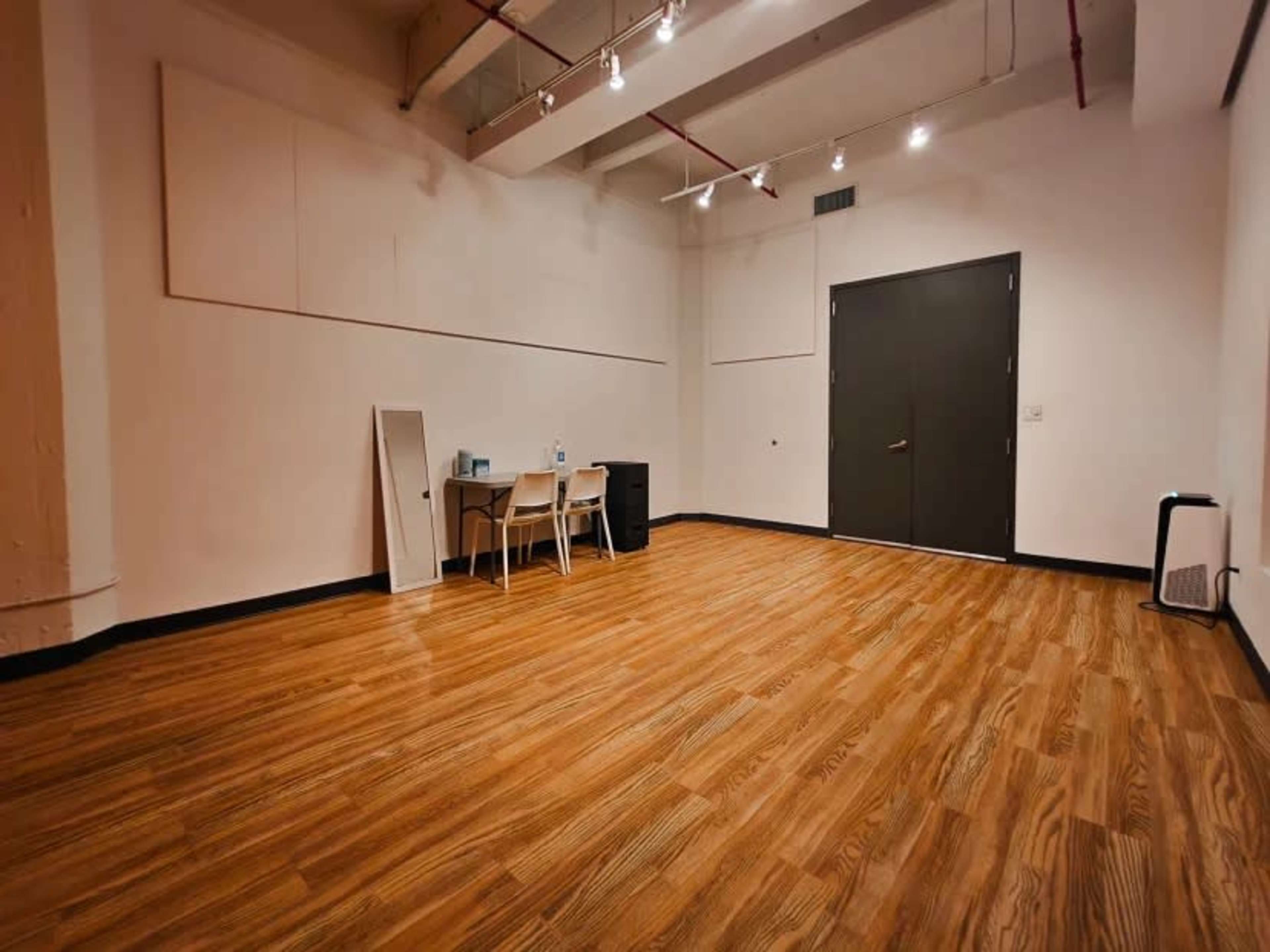 The image shows a sparsely furnished room with wooden flooring, featuring a table with chairs, a mirror, and a door on one side.