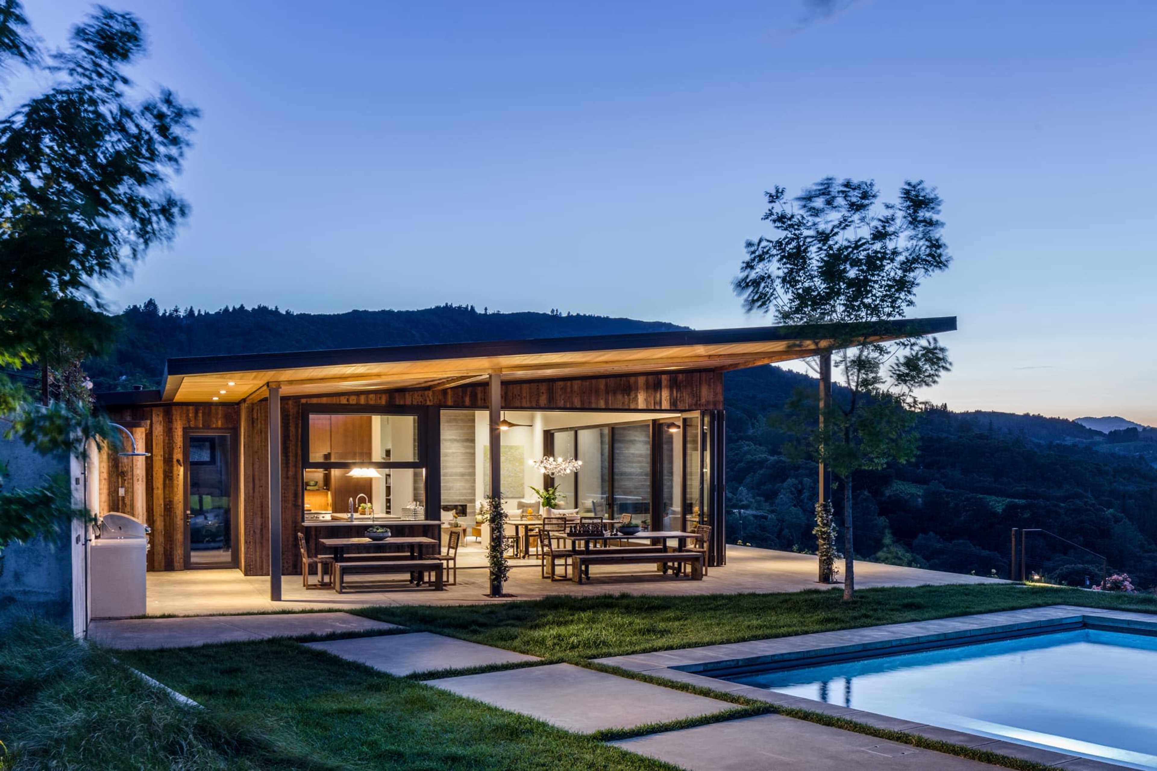 A modern wooden house with large glass windows overlooks a pool and landscaped area at twilight.