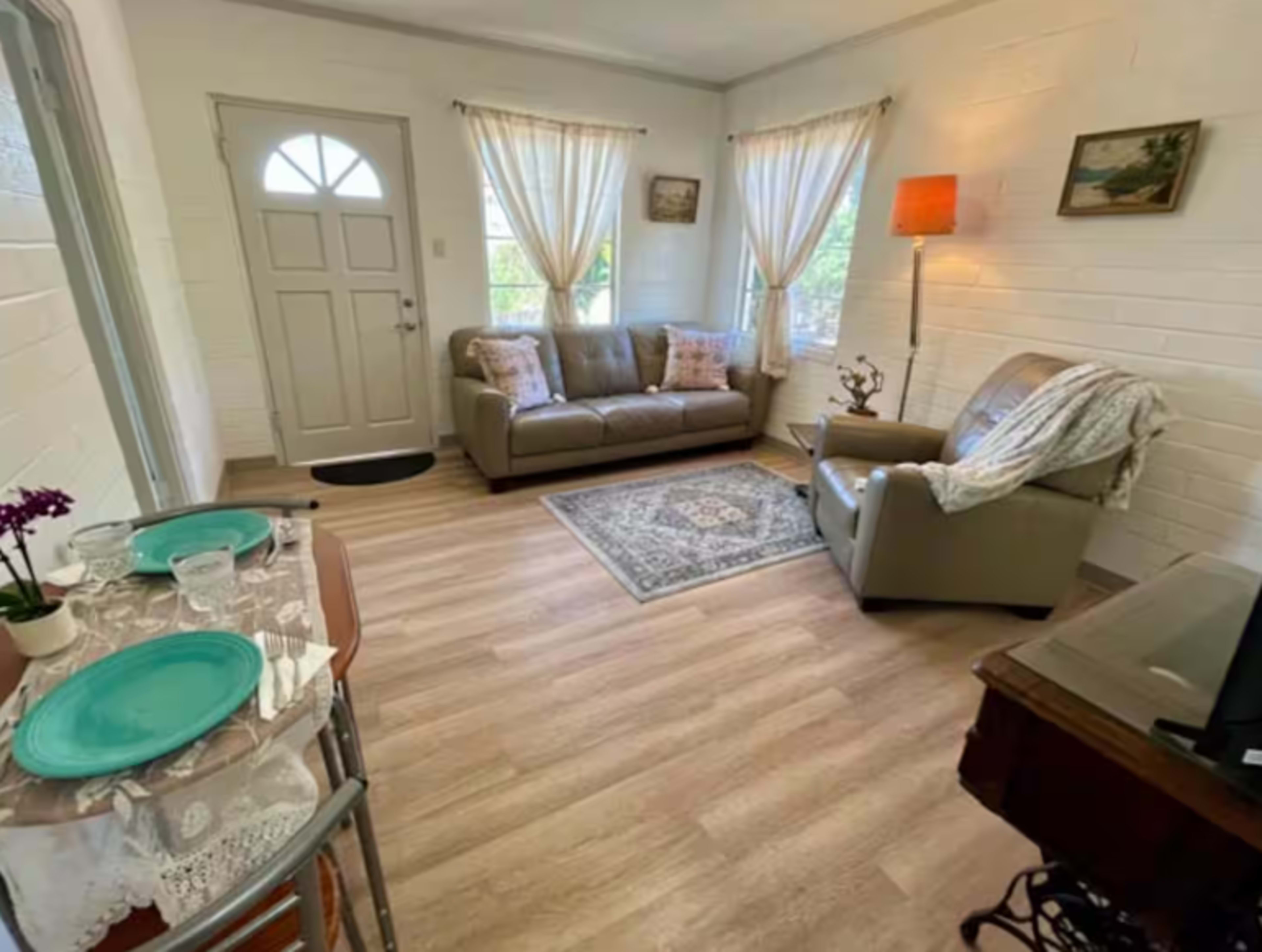 The image shows a small living room with a sofa, an armchair, a small dining table set for two, and a door leading outside.