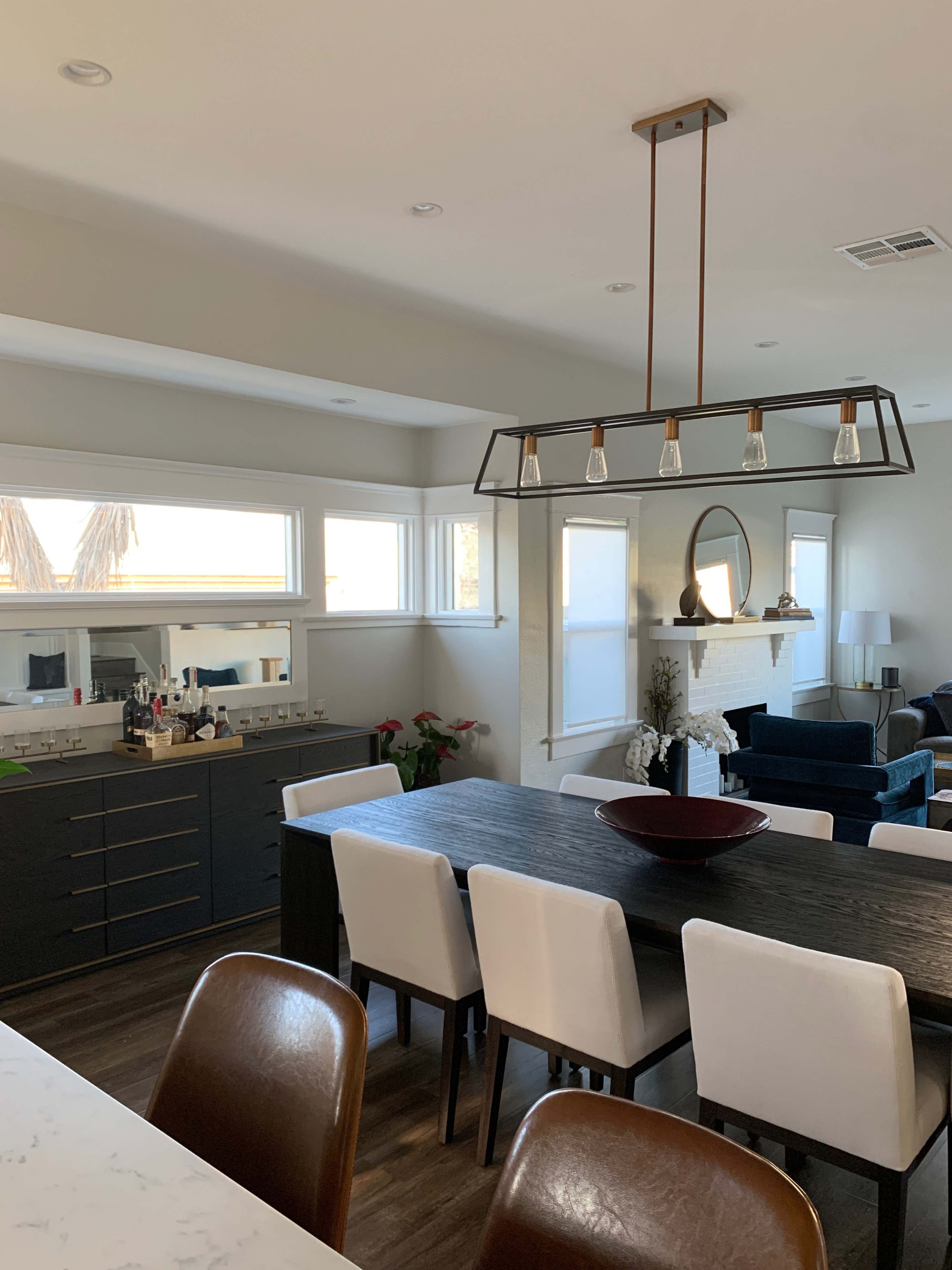 The image shows a modern dining area with a long wooden table surrounded by white and brown chairs, a chandelier above, and large windows letting in natural light.
