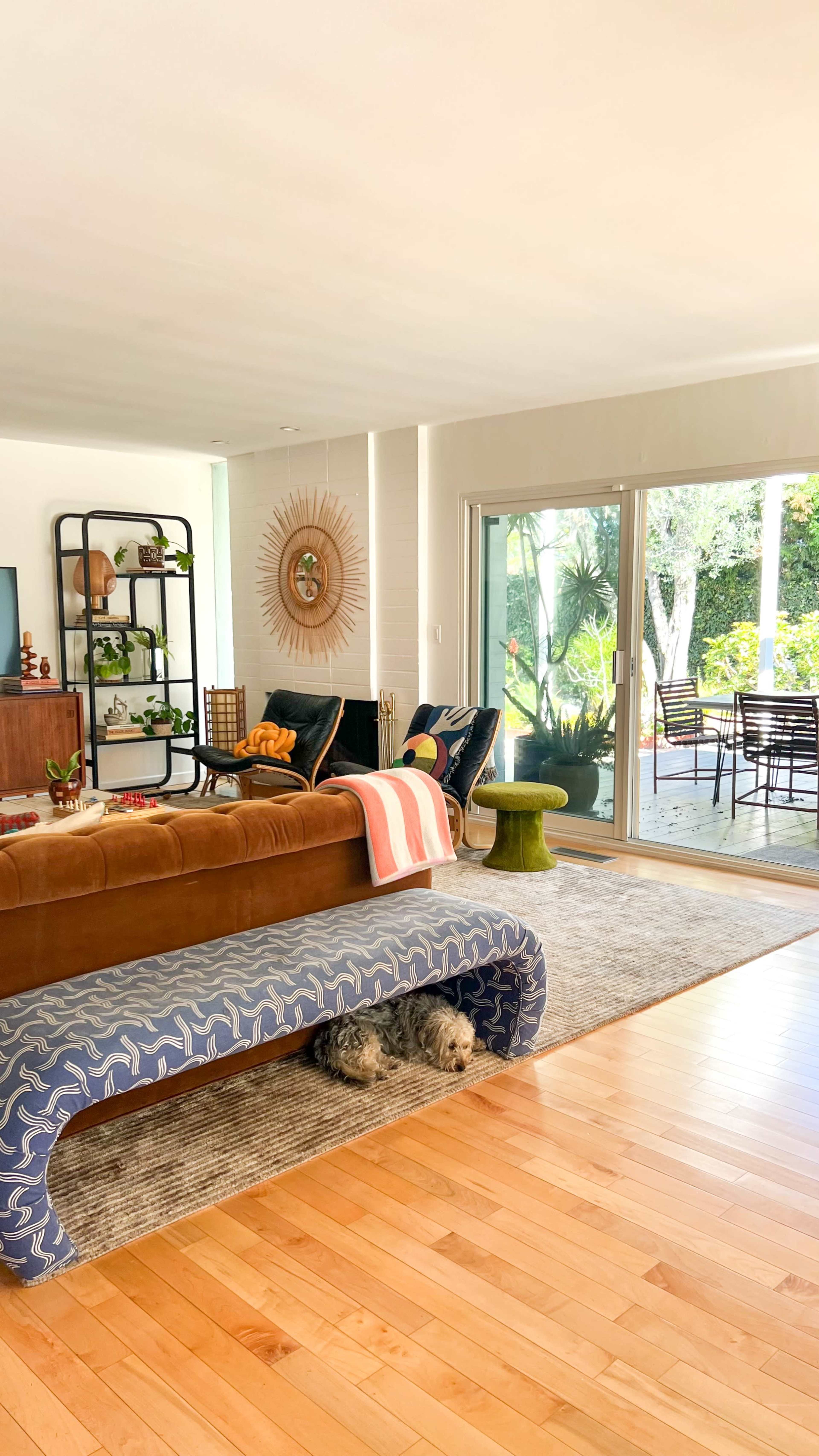The image shows a contemporary living room with a brown sofa, a patterned coffee table, and large windows overlooking a patio area with outdoor seating.