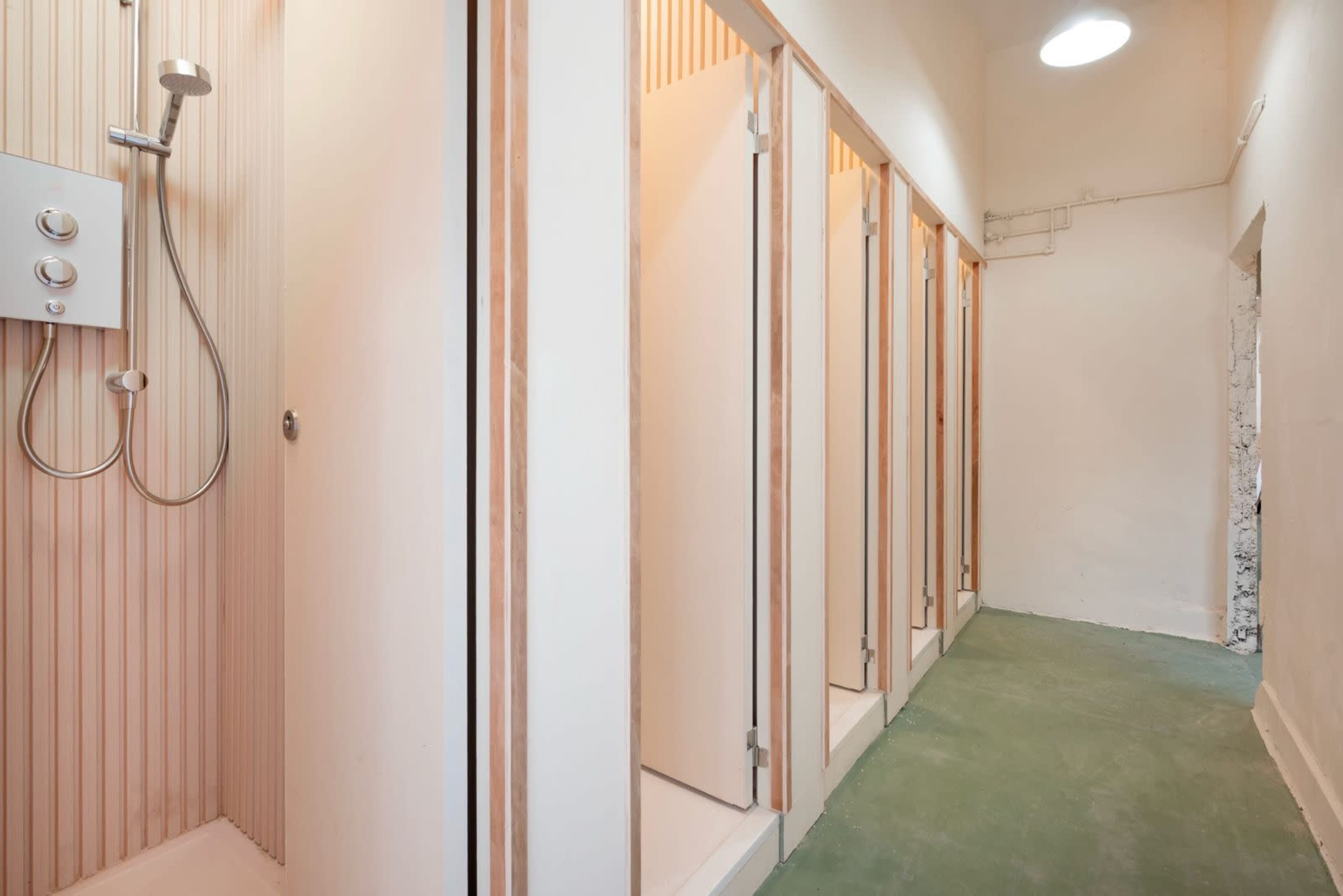 The image shows a row of shower stalls with wooden doors and a shower installation on the left side of a minimalist, modern bathroom.