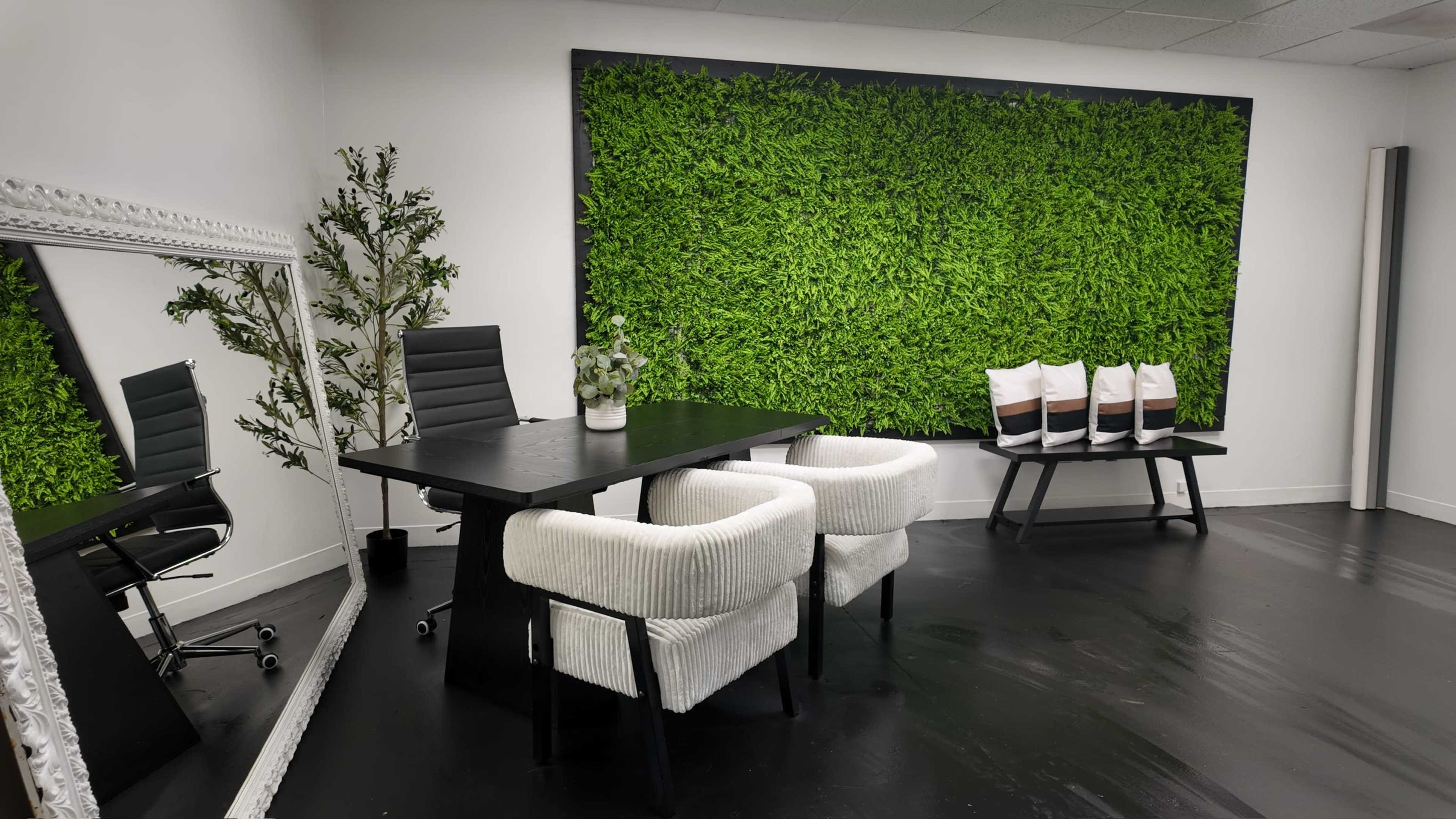 The image depicts a modern office space featuring a black table with two white upholstered chairs, a large mirror, a potted plant, and a green wall covered in artificial foliage.