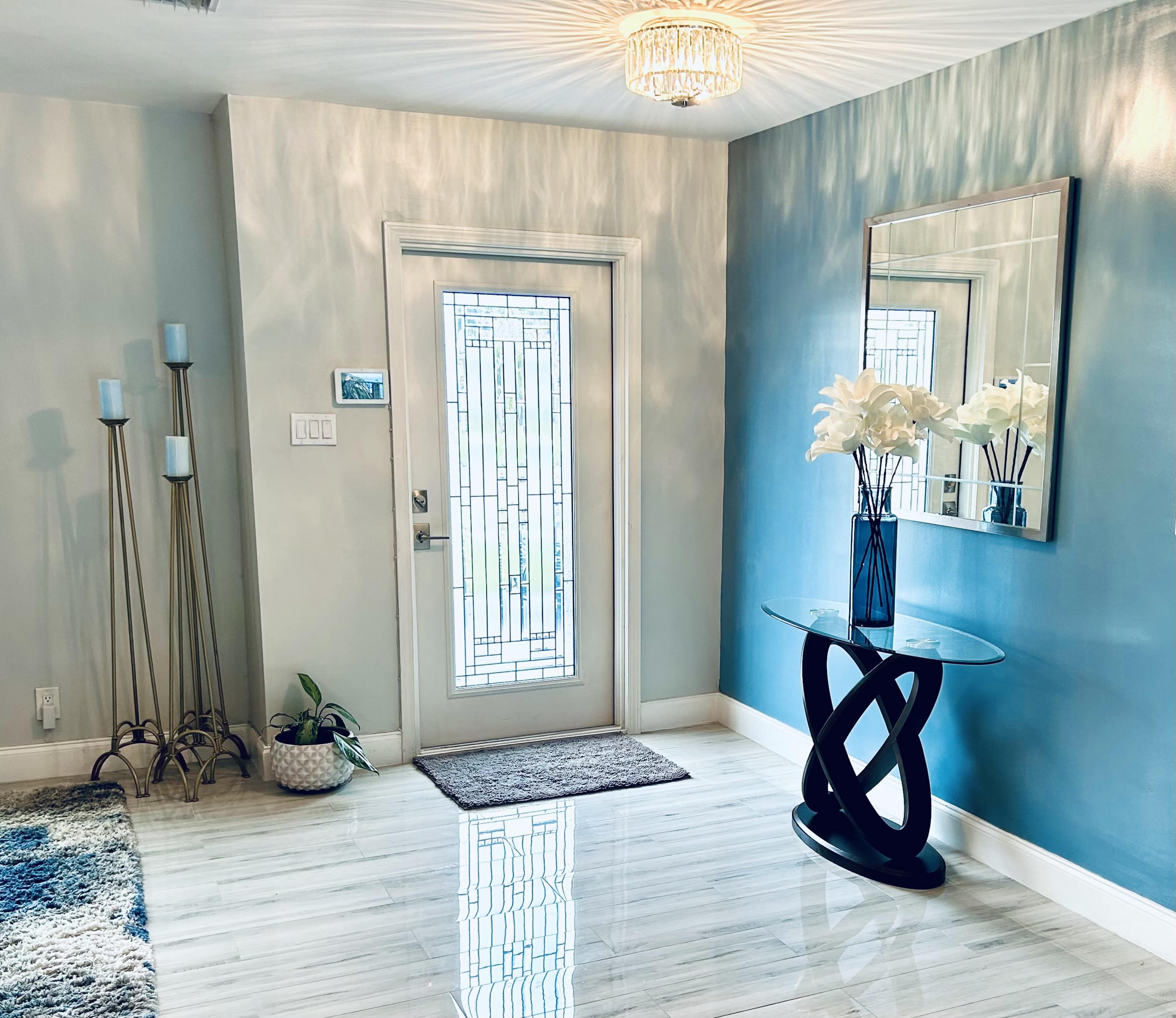 A modern entryway featuring a decorative glass door, a round glass table with an abstract black base, and a wall-mounted mirror, complemented by a vase of white flowers and candle holders.