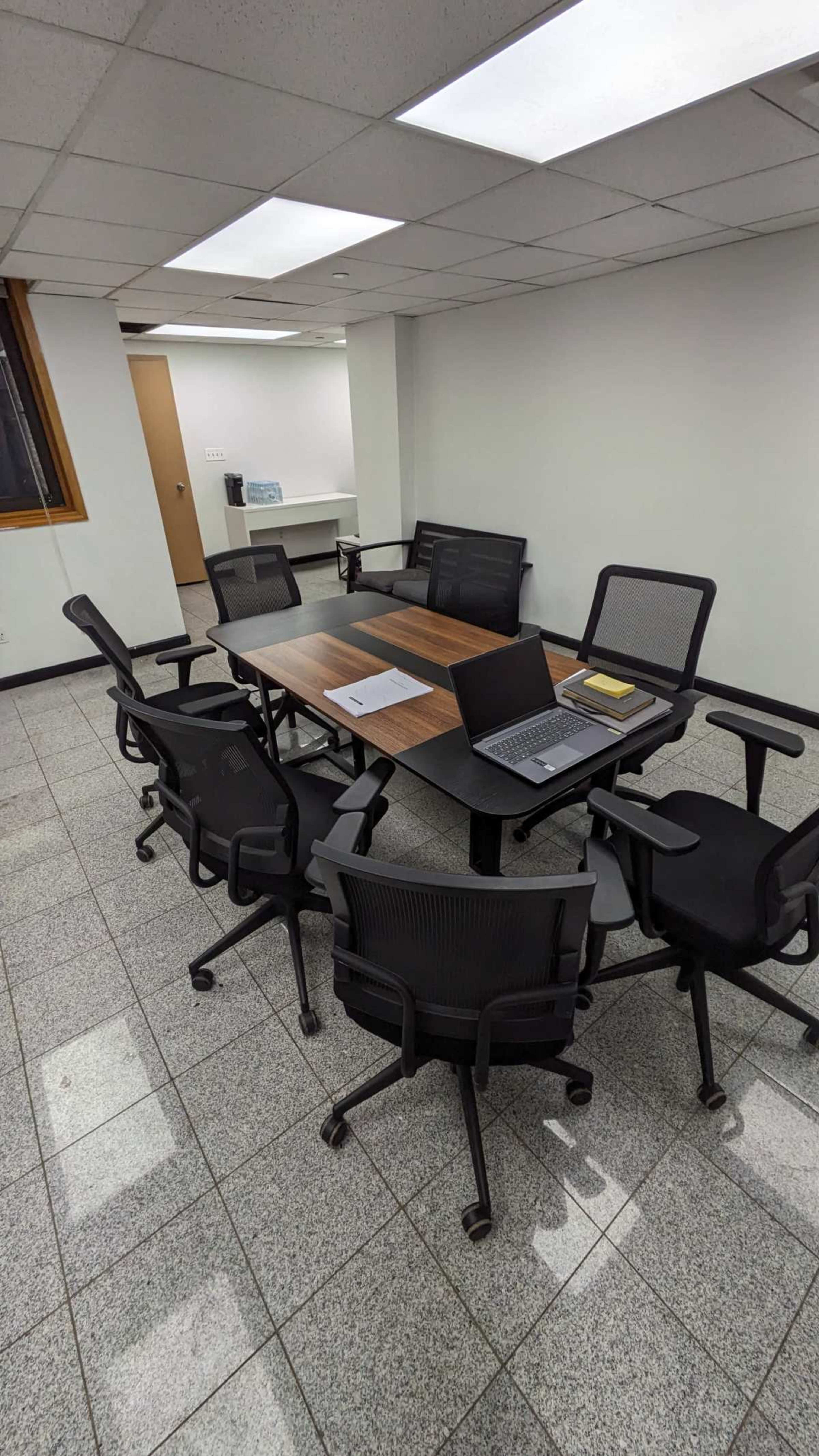 A meeting room features a large wooden table surrounded by six office chairs, with a laptop and papers on the table.