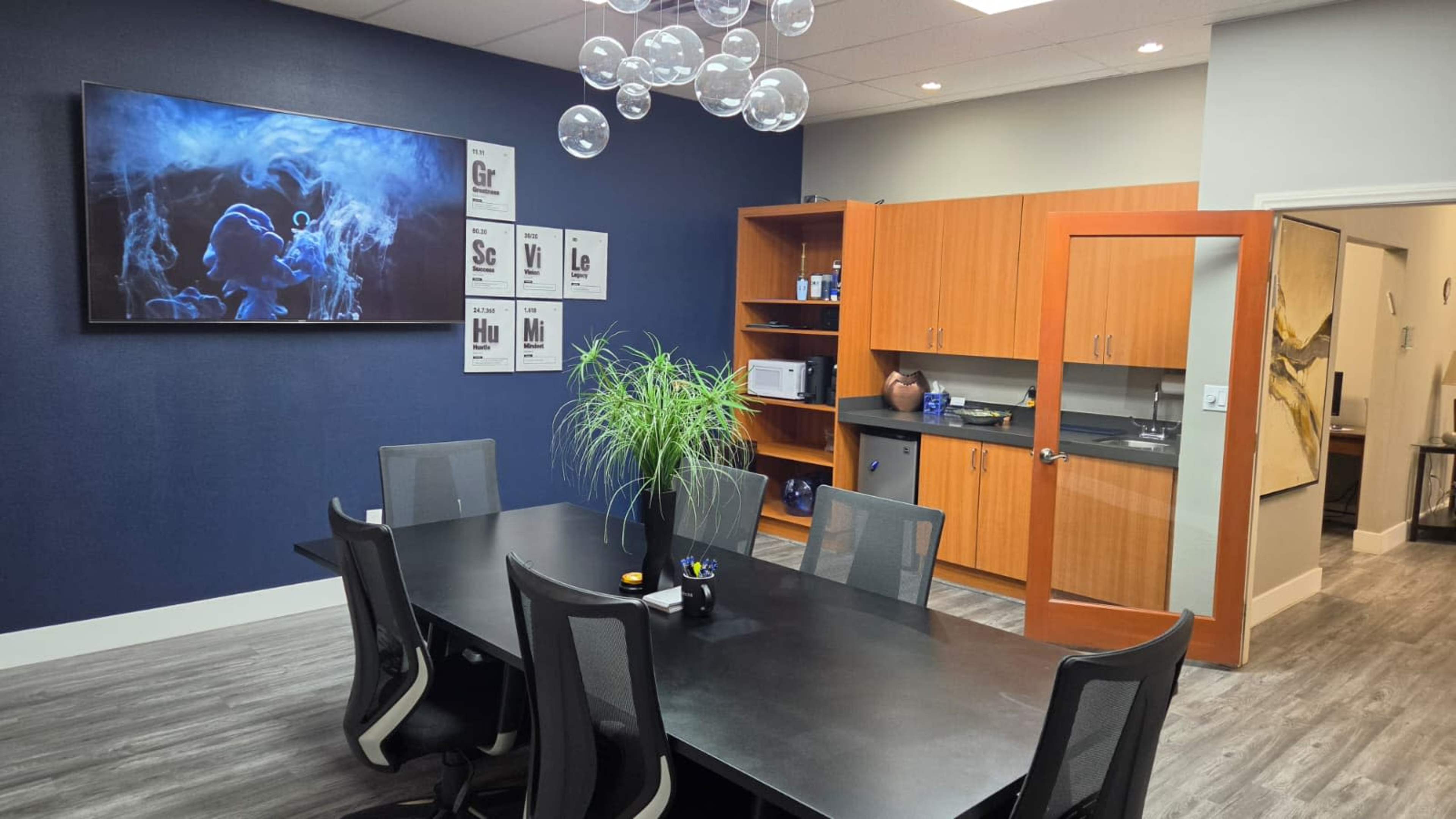 The image shows a modern conference room with a long black table, ergonomic chairs, a large television mounted on the wall, and cabinetry containing various office supplies.