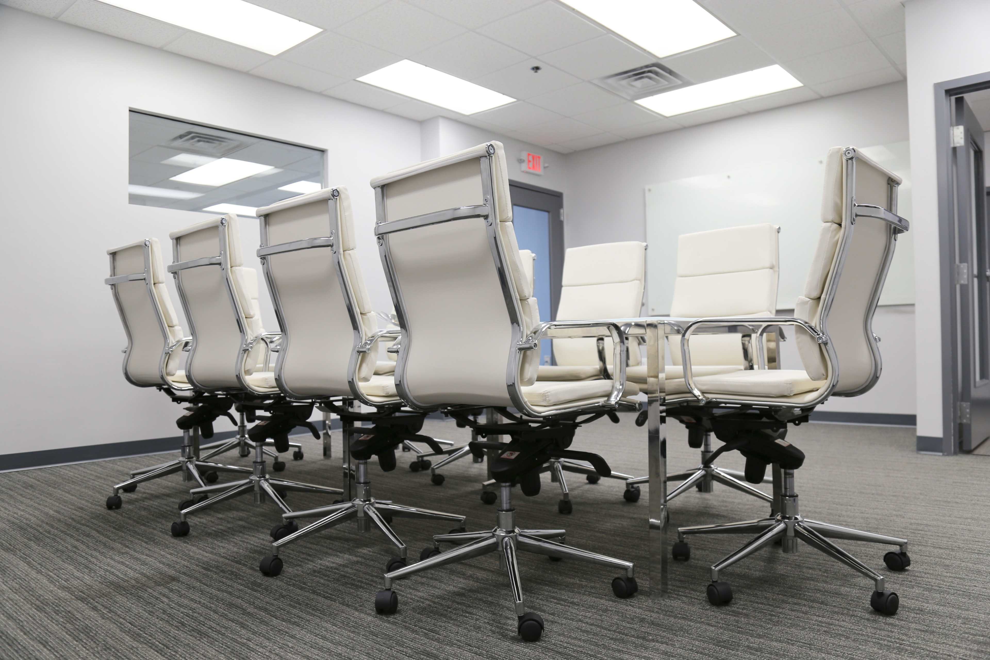 A row of eight white office chairs on wheels is positioned around a meeting room table in a modern workspace.