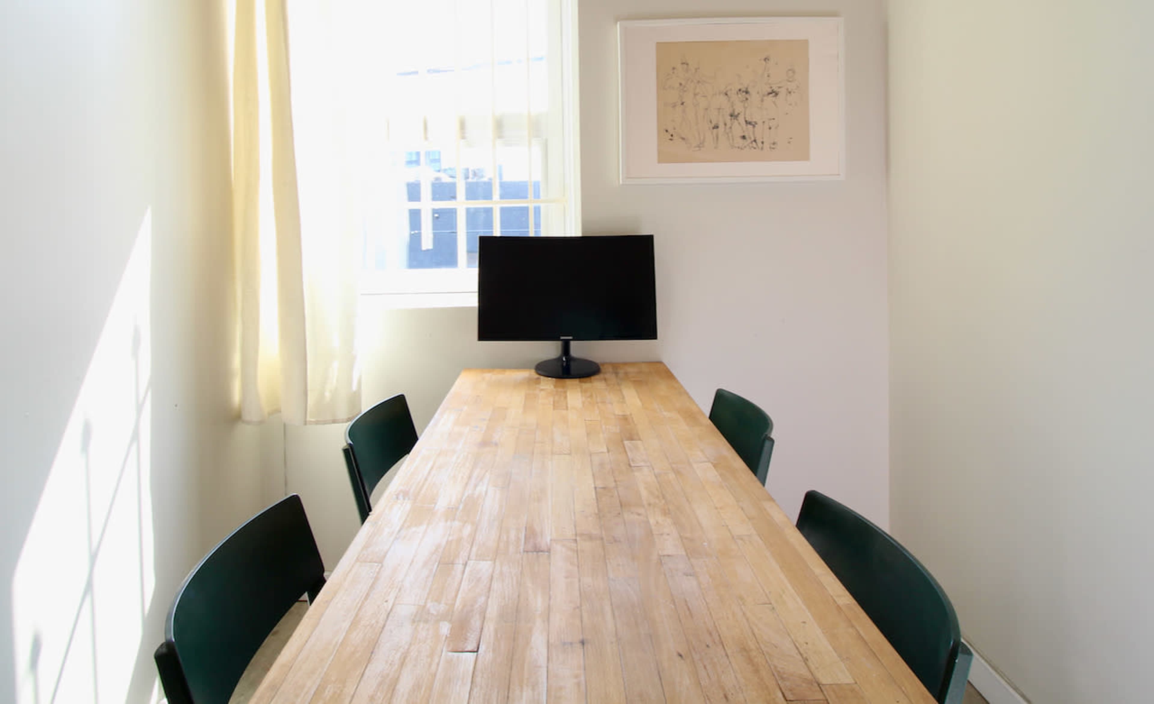 A bright, minimalist meeting room features a wooden table with four green chairs and a monitor on a stand at one end.
