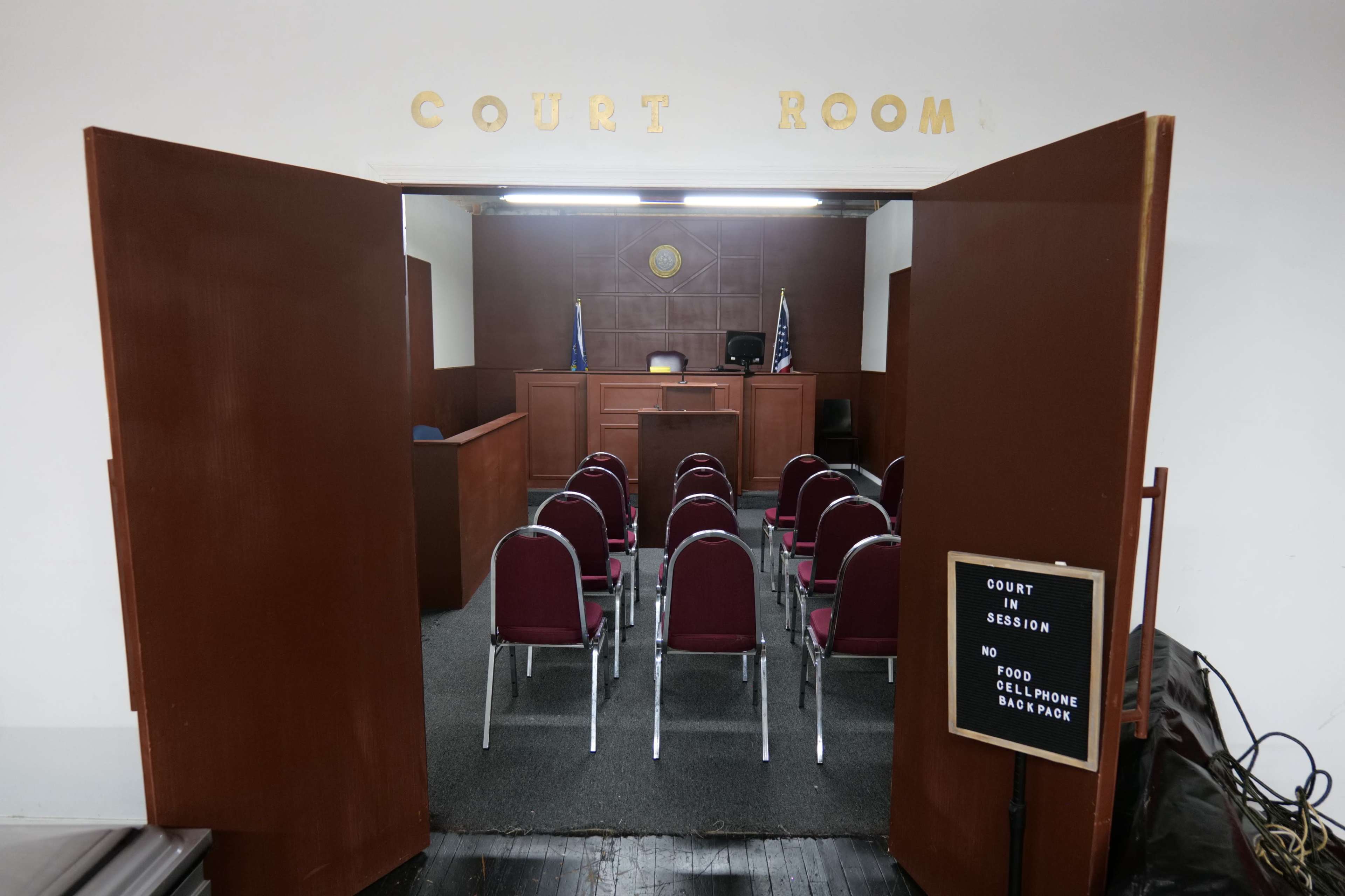 A courtroom with wooden doors partially open, revealing empty rows of chairs and a judge's bench at the front.