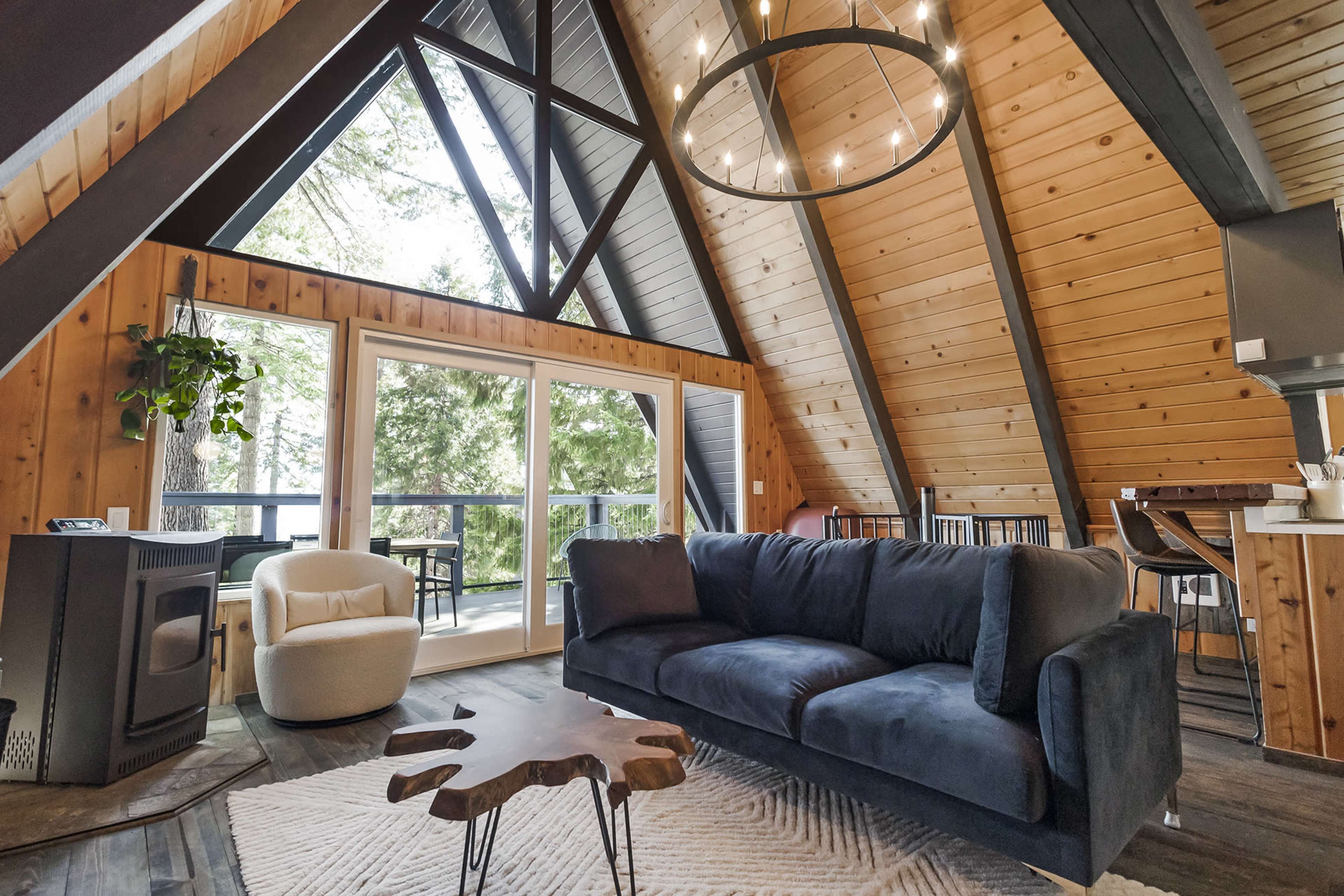 The interior of a modern A-frame cabin features a large window, wooden walls, and a cozy seating area with a circular light fixture overhead.