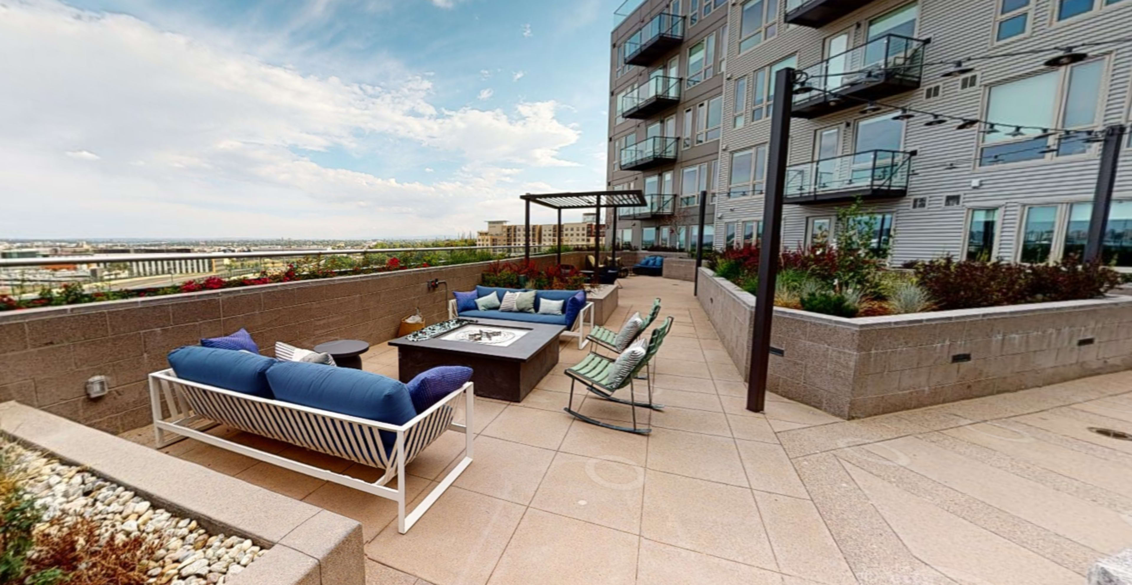 A rooftop patio area featuring a seating arrangement with a table, lounge chairs, and surrounding greenery, adjacent to a multi-story residential building.
