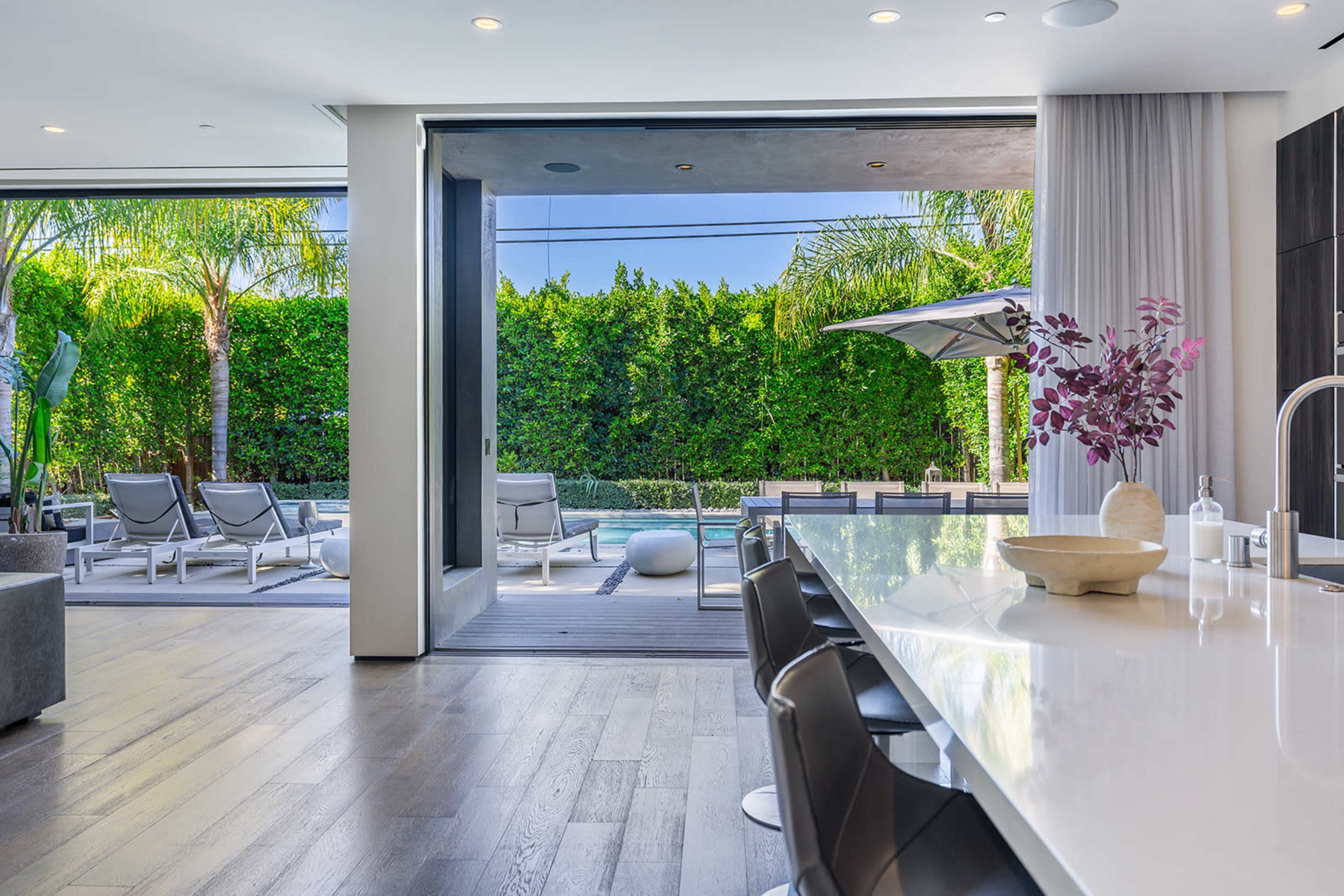 The image shows a modern kitchen area overlooking a pool with sun loungers and greenery in the background.