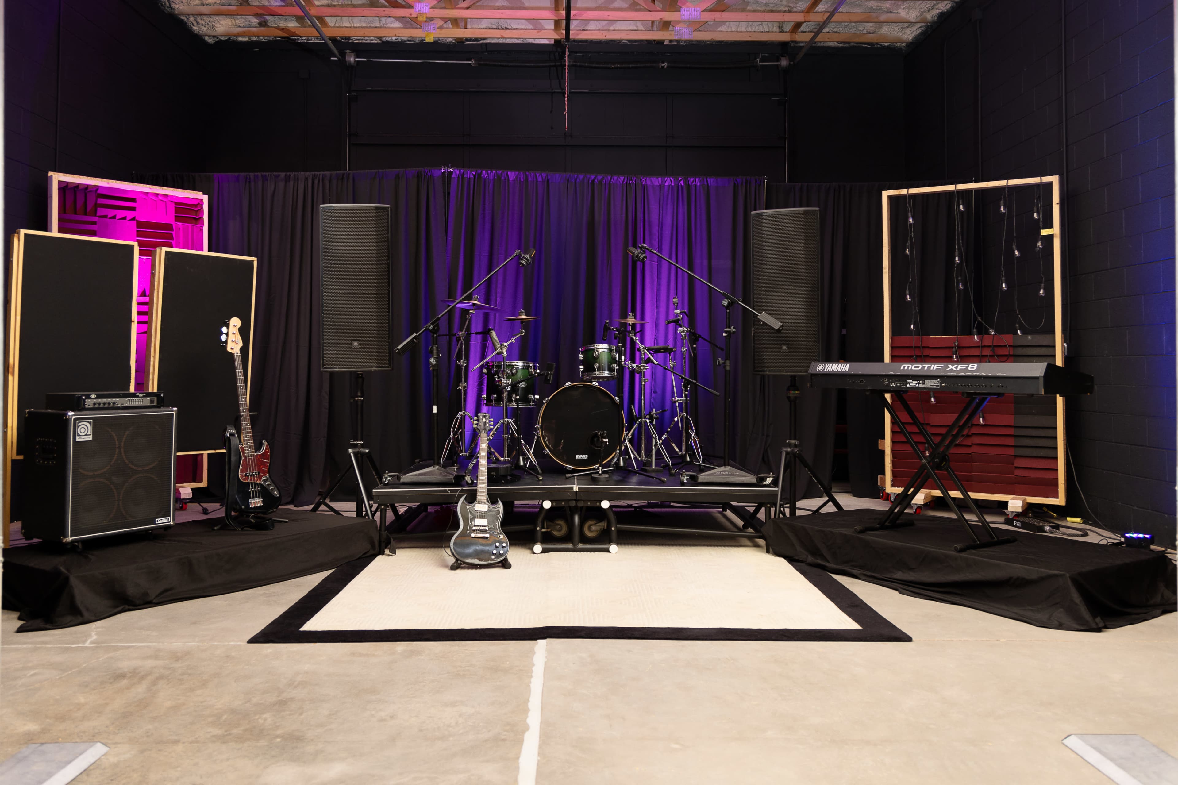 A stage setup features a drum kit, keyboard, electric guitar, amplifiers, and speakers, with black curtains and purple lighting in the background.