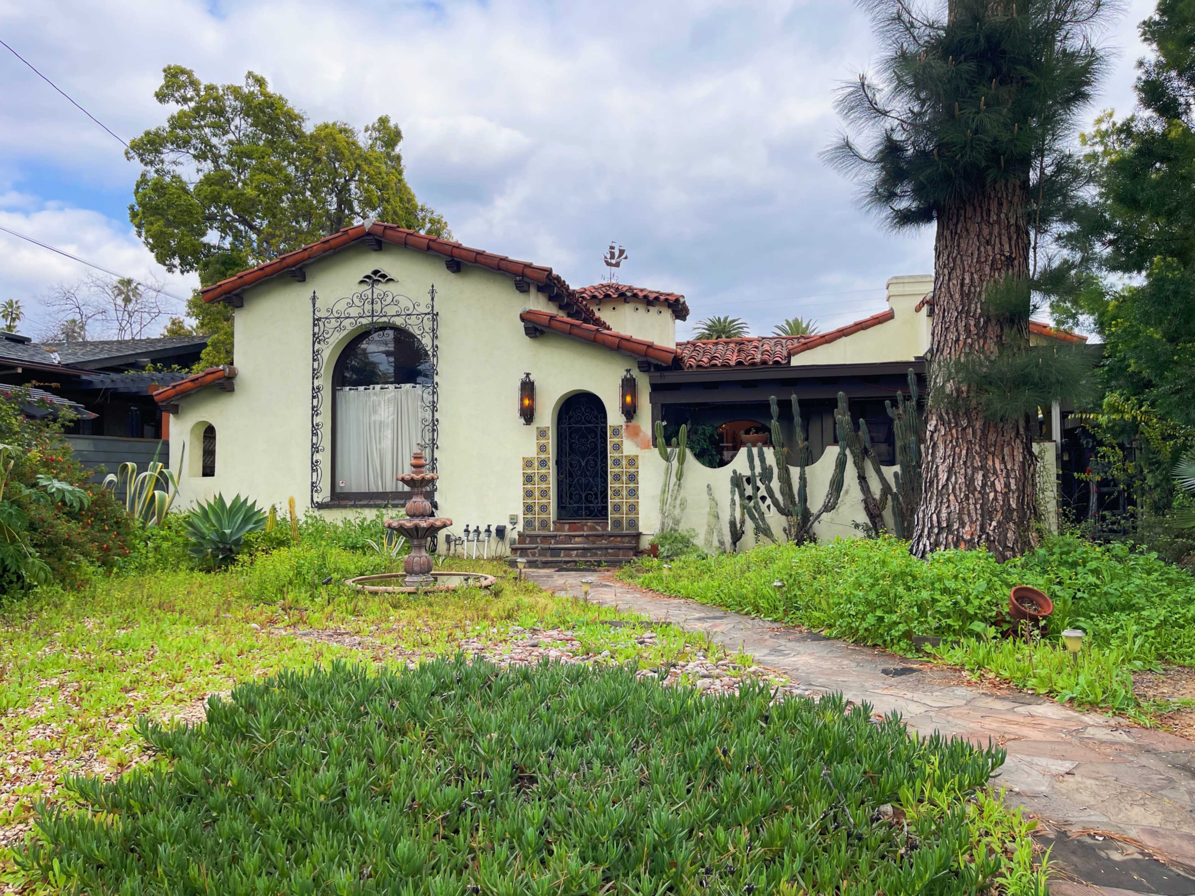 A stucco house with a terracotta roof, a front garden featuring green plants, and a stone pathway leading to the entrance.