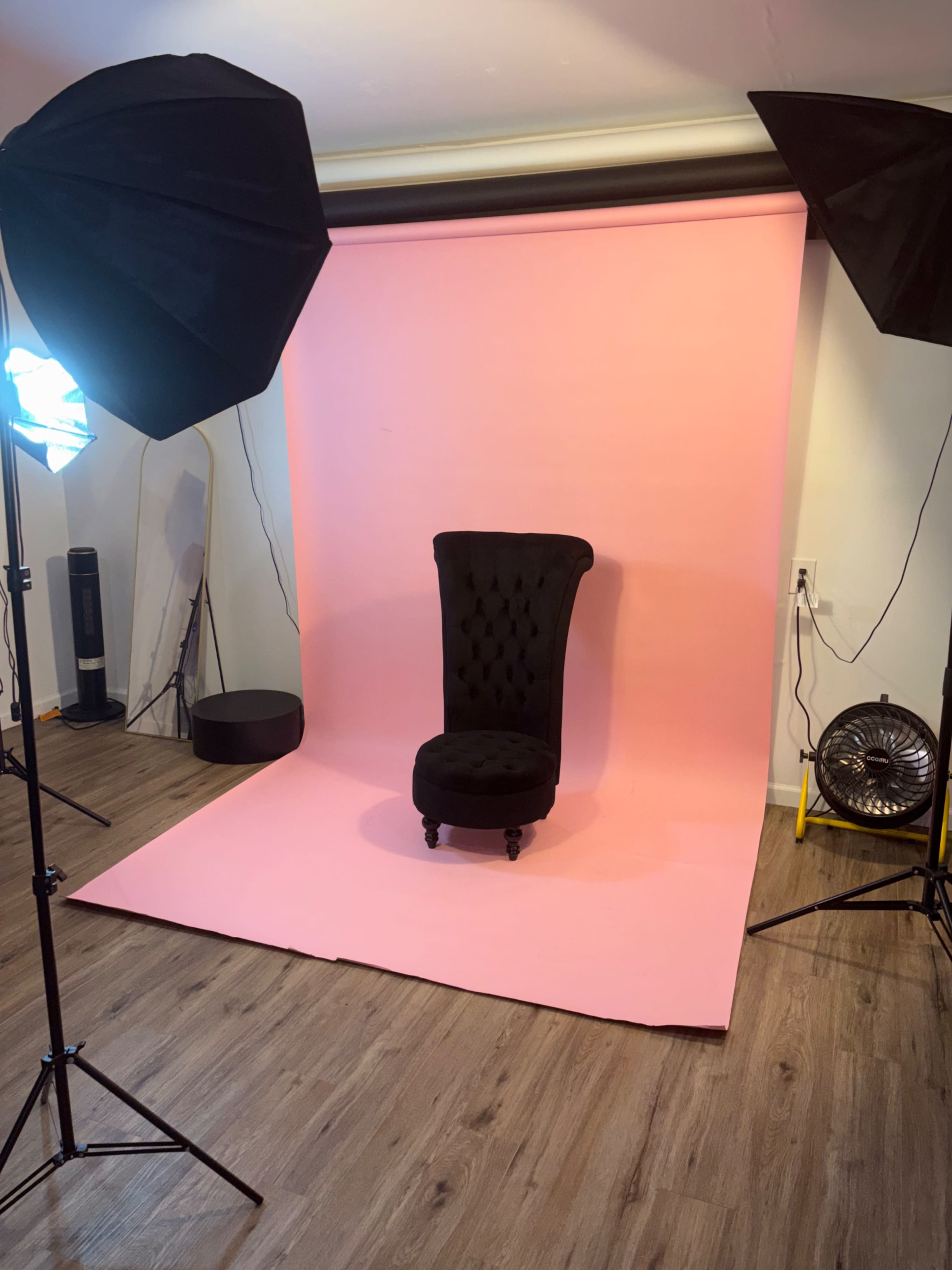 The image shows a photography studio setup with a black tufted chair in front of a pink backdrop, surrounded by lighting equipment.