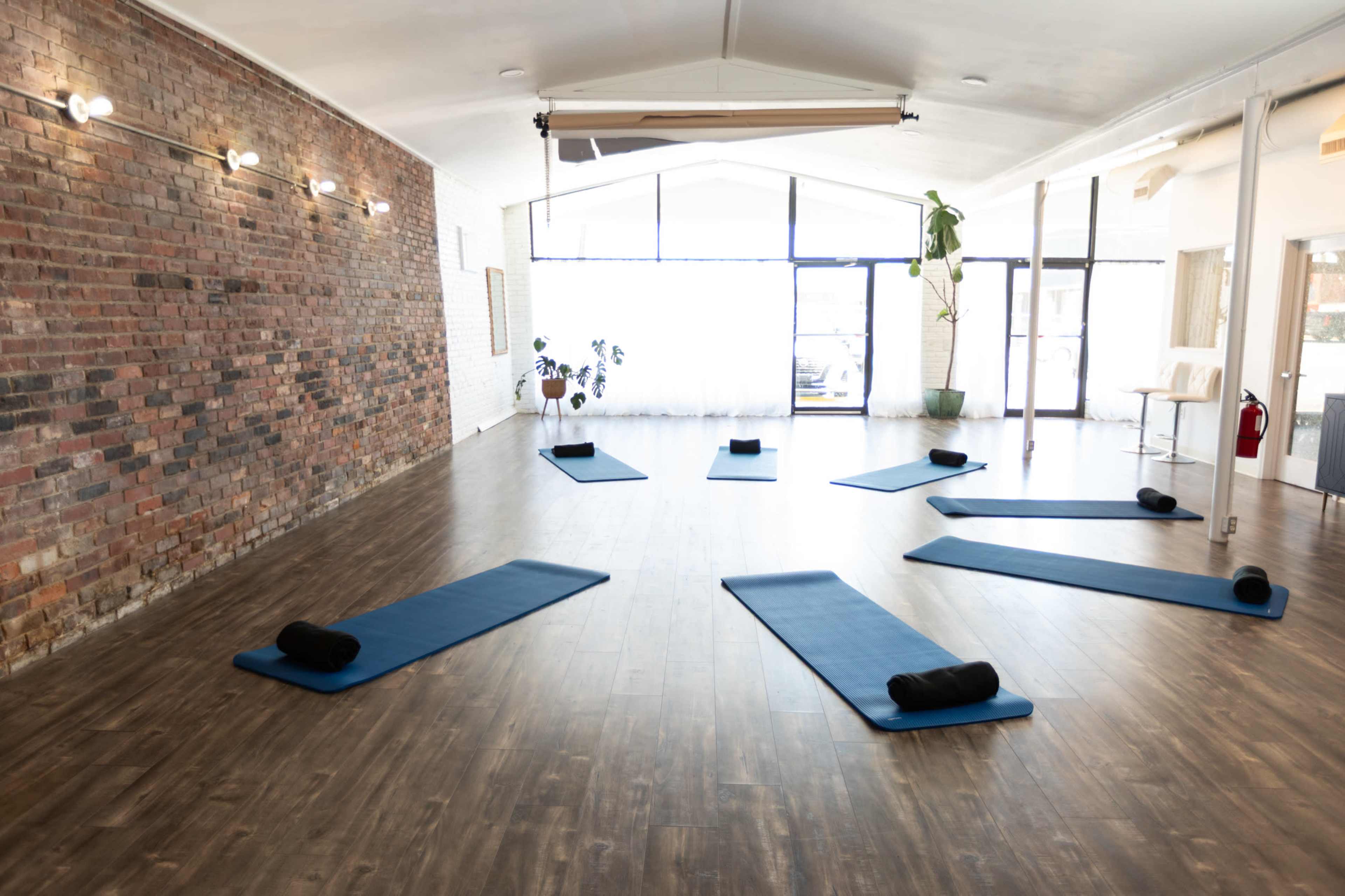 The image shows a spacious yoga studio with several blue mats arranged on a wooden floor, alongside a brick wall and large windows letting in natural light.