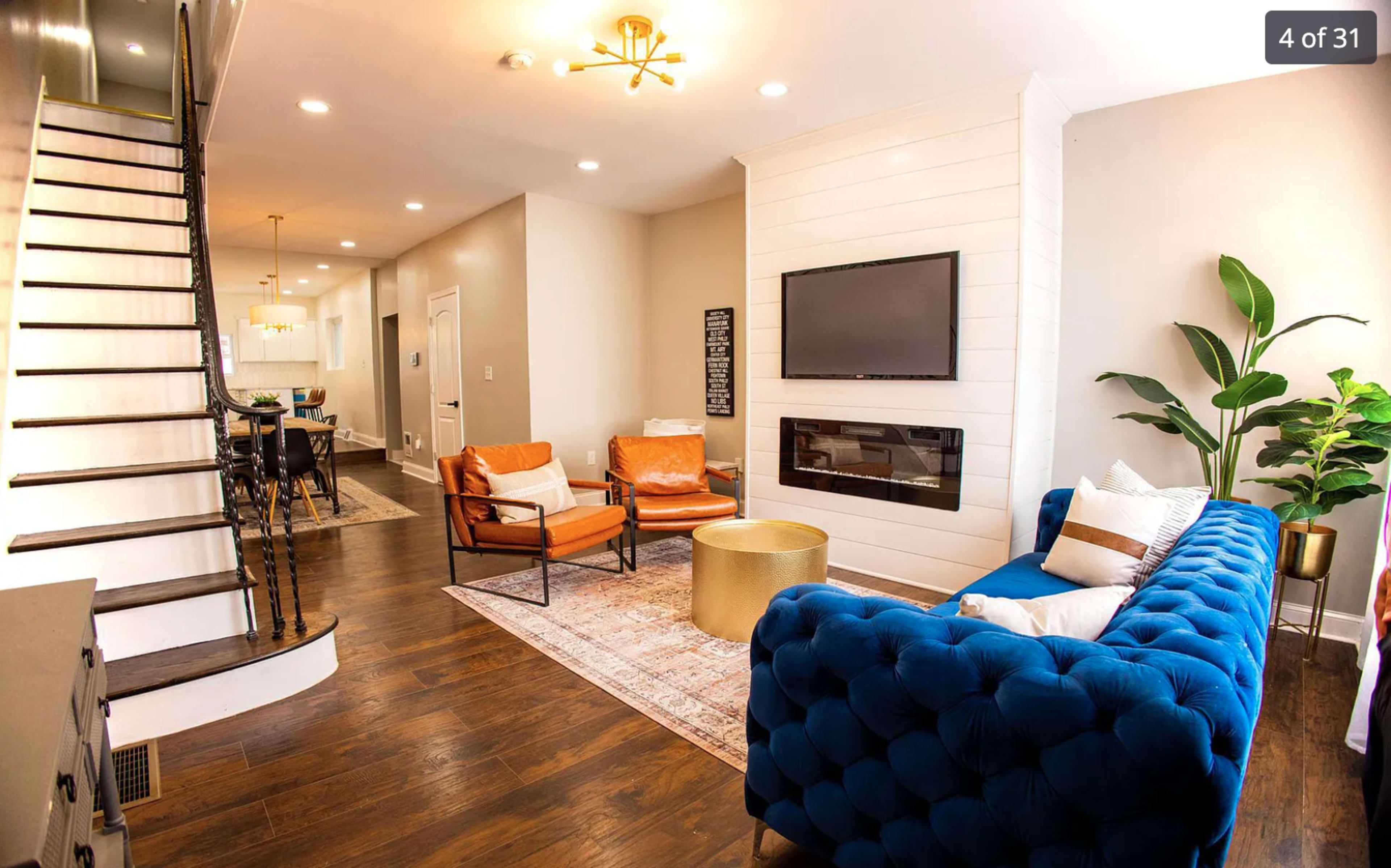 A modern living room with a blue tufted sofa, two orange armchairs, a gold coffee table, a mounted TV, and a staircase in the background.