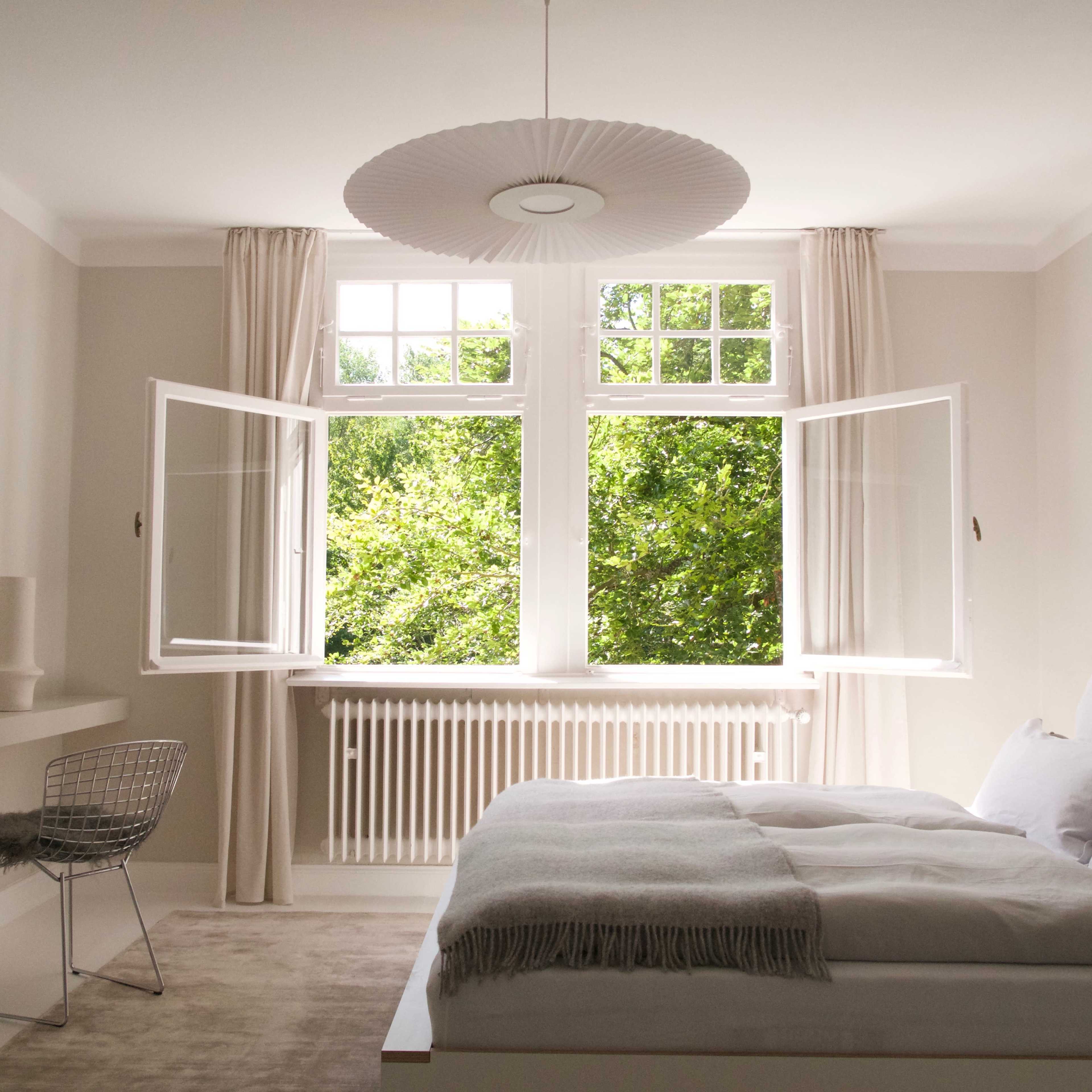 A minimalist bedroom features a bed with white linens, a large window with open doors overlooking greenery, and simple décor.