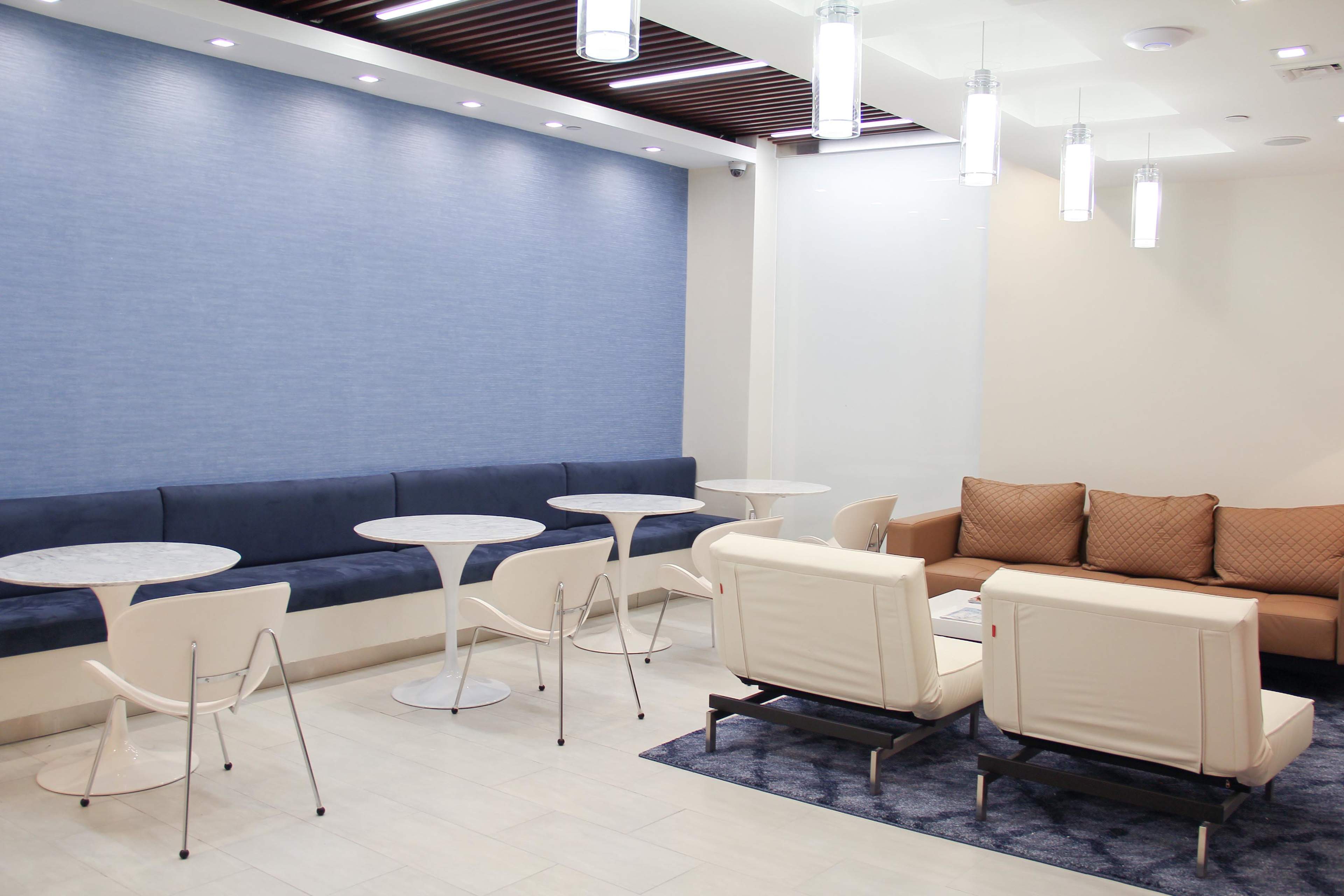 The image shows a modern lounge area featuring a blue accent wall, several small round tables, and white chairs arranged around a seating area with brown cushions.