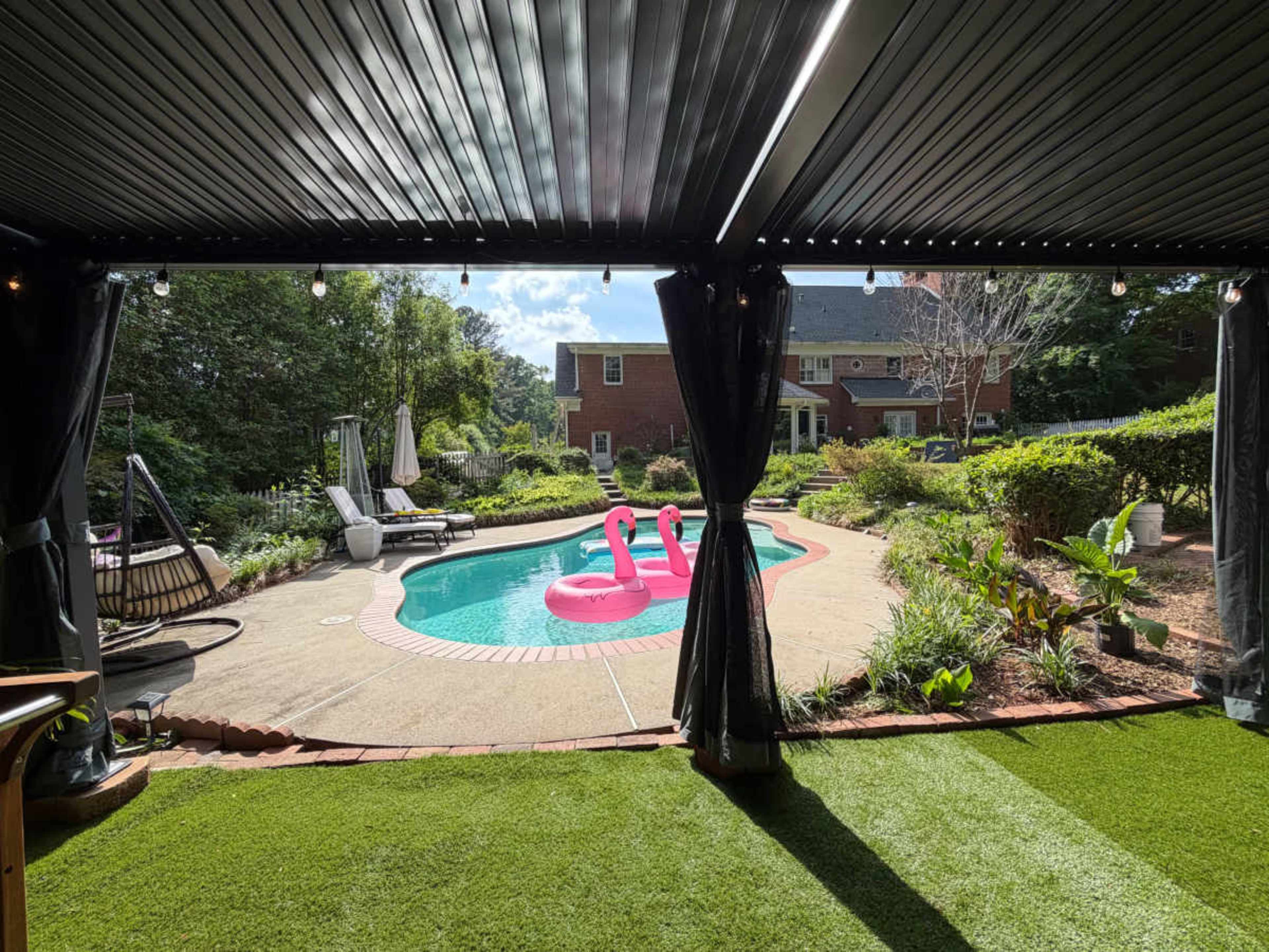The image shows a backyard pool area with two pink flamingo floaties, surrounded by a well-manicured garden and patio furniture.