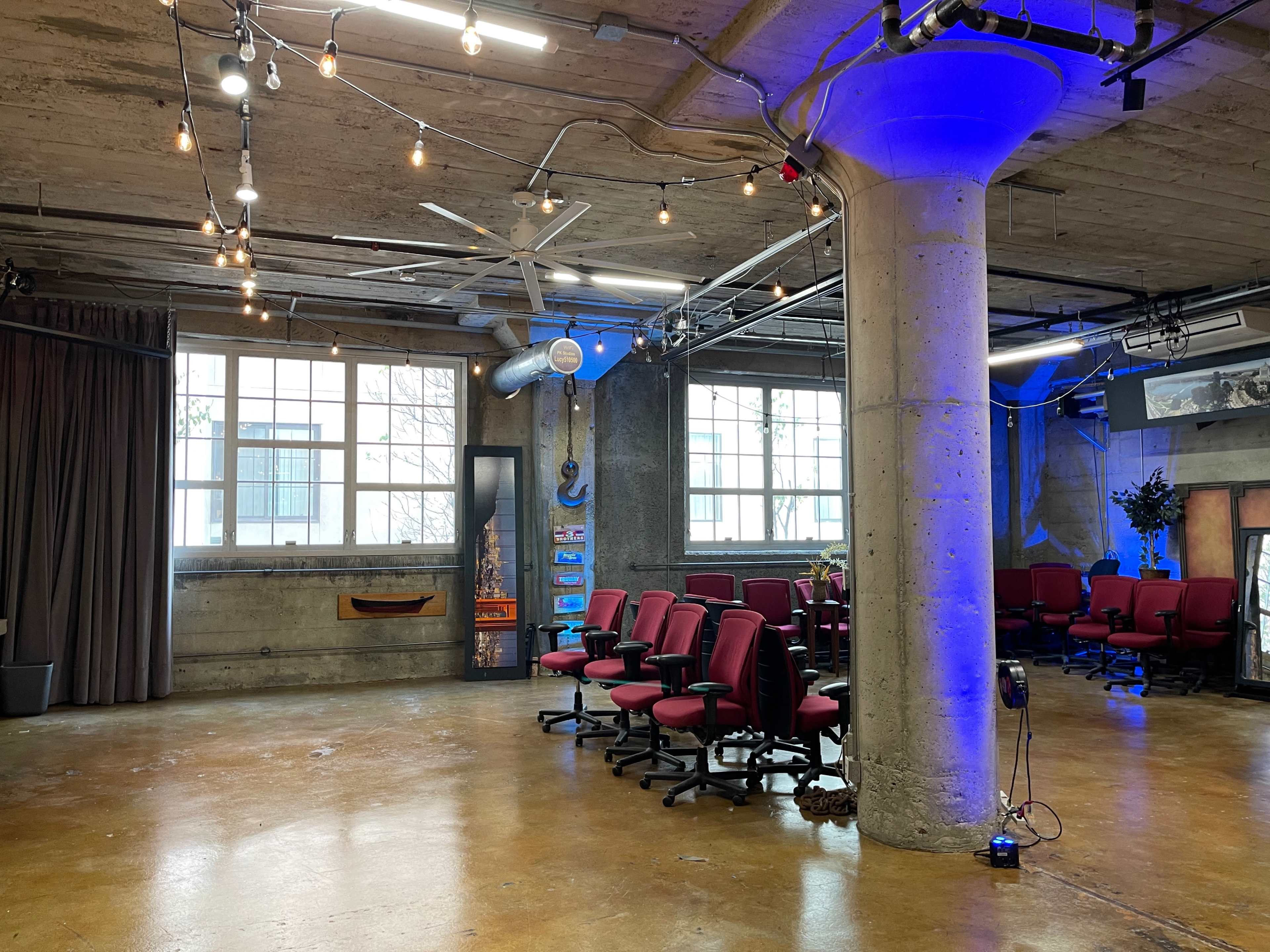 A spacious, industrial-style meeting room with rows of red office chairs, exposed concrete walls, and large windows.