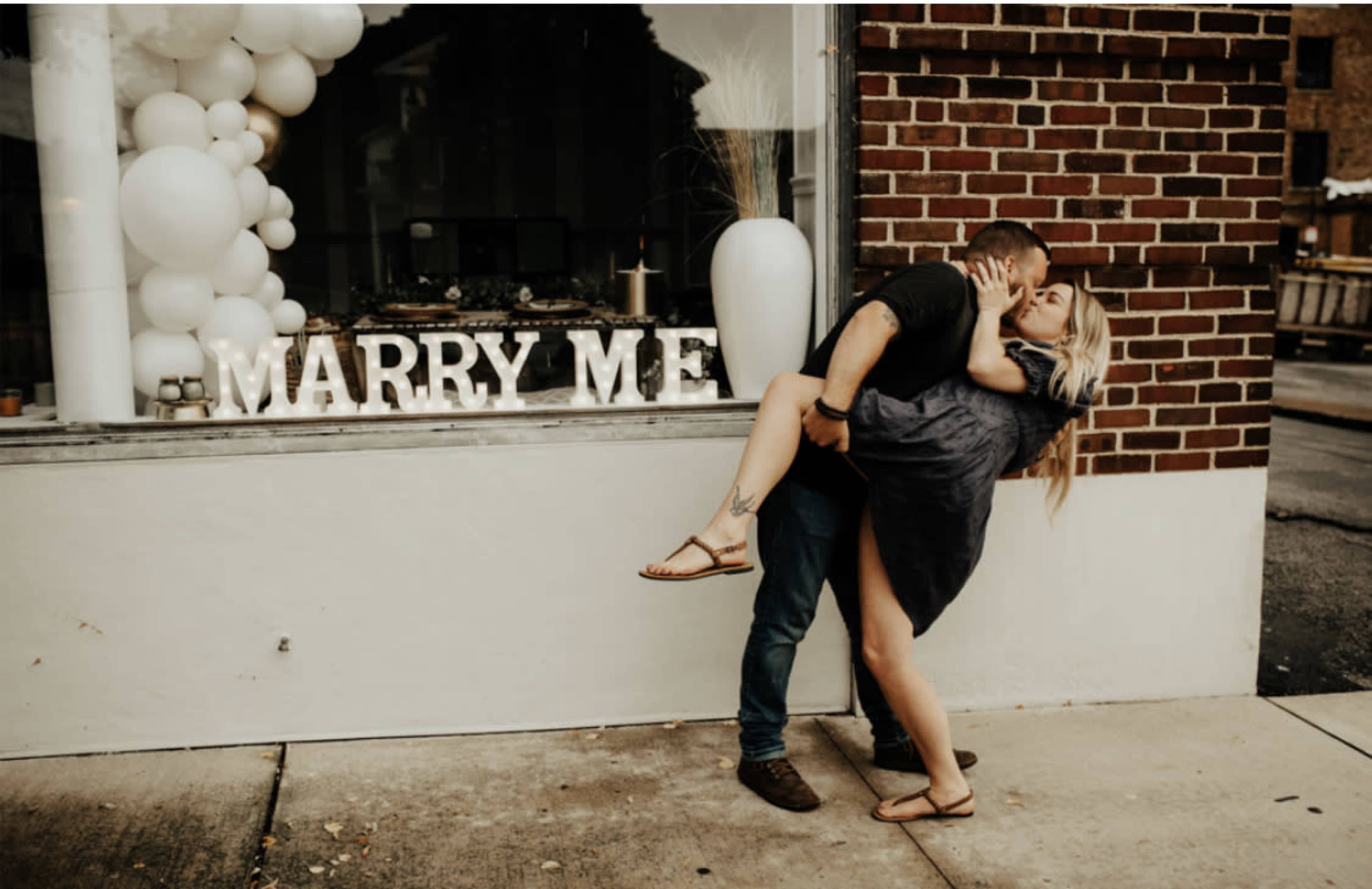 A couple embraces passionately outside a store featuring a "MARRY ME" display.