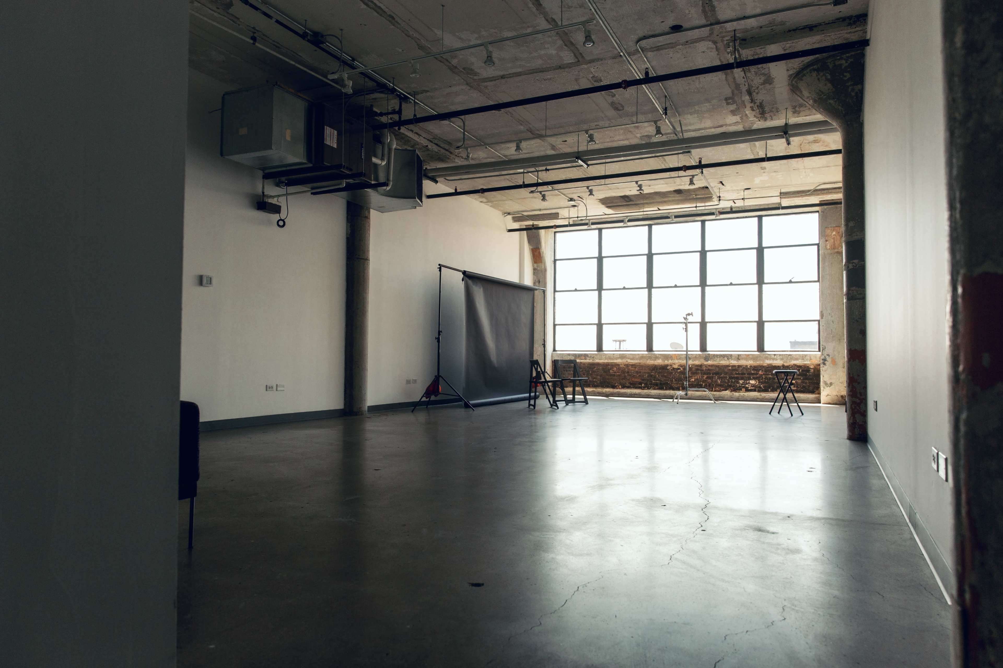 An empty industrial-style room with exposed concrete walls, large windows, and a backdrop stand at one end.