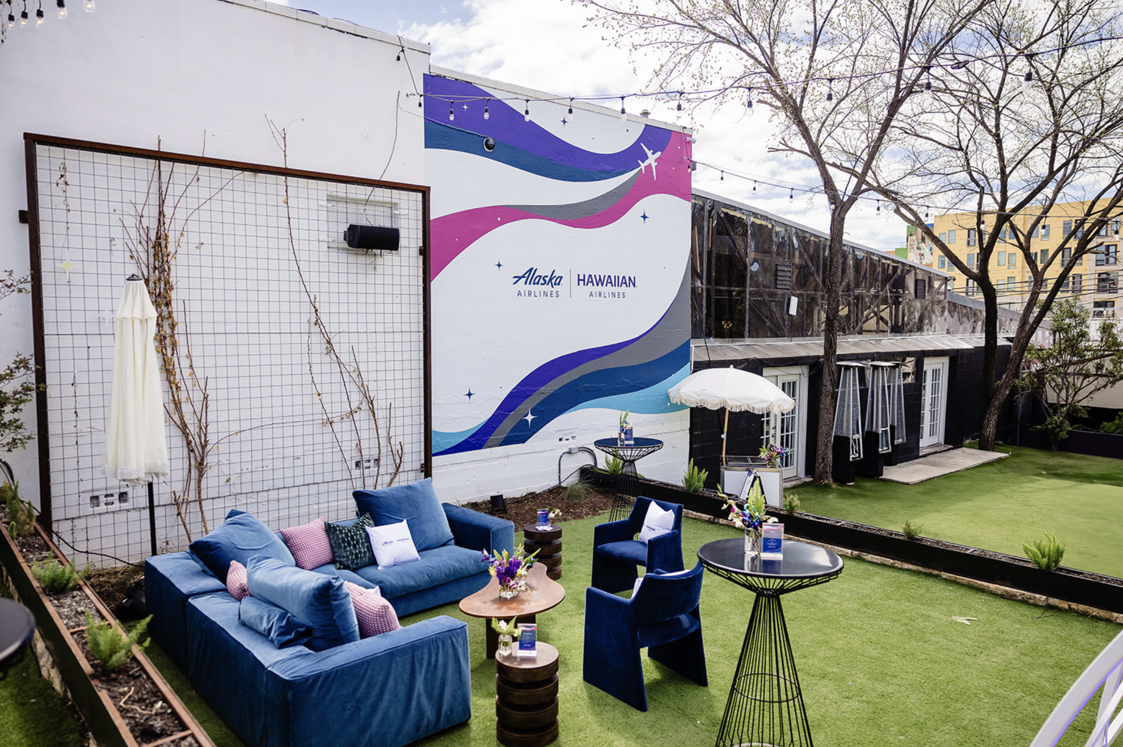A vibrant outdoor lounge area features blue sofas, a decorative table, and a colorful wall mural with the logos of Alaska Airlines and Hawaiian Airlines.