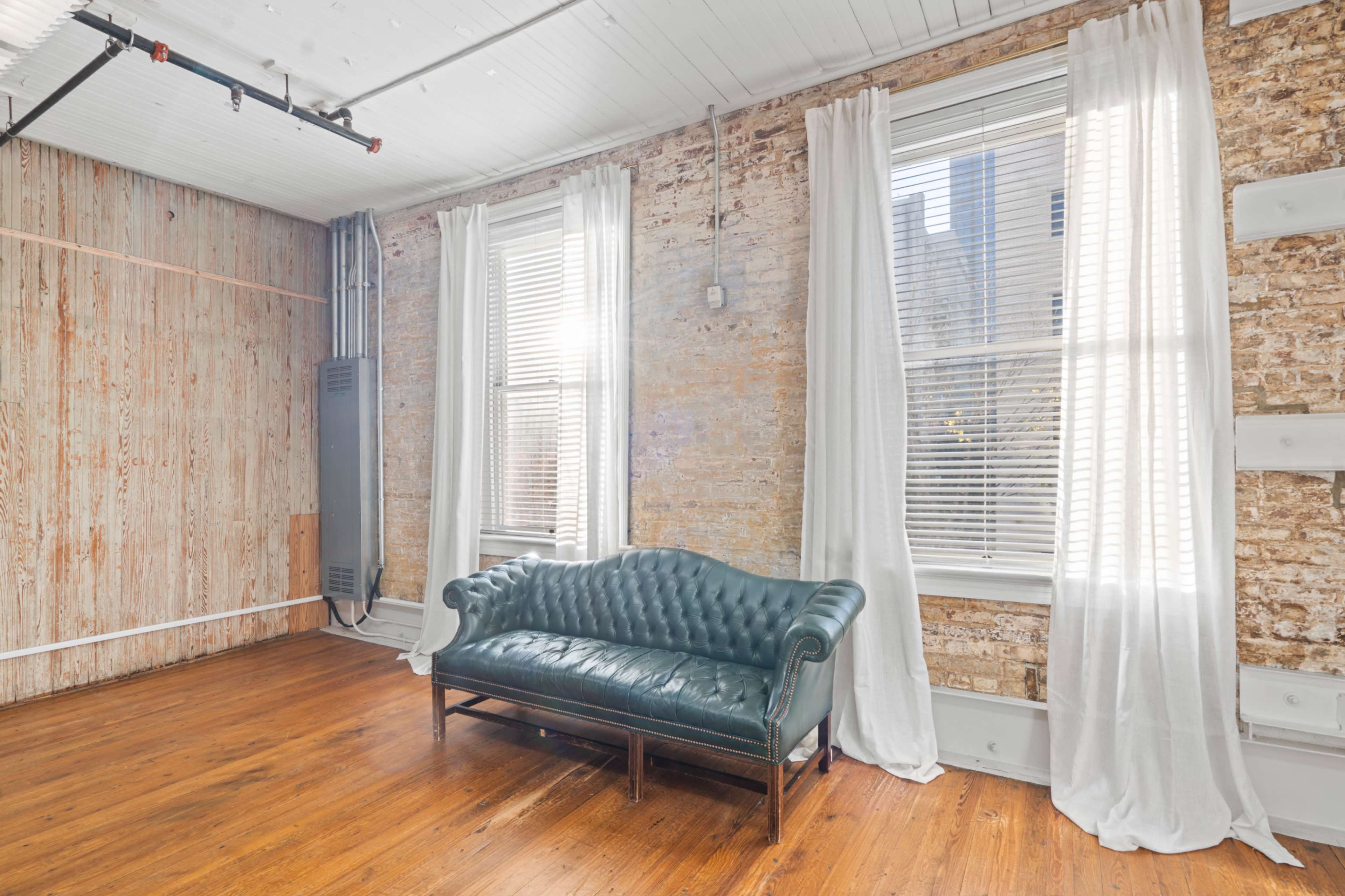 The image shows a room with exposed brick walls, two windows with white curtains, and a green leather sofa on wooden flooring.