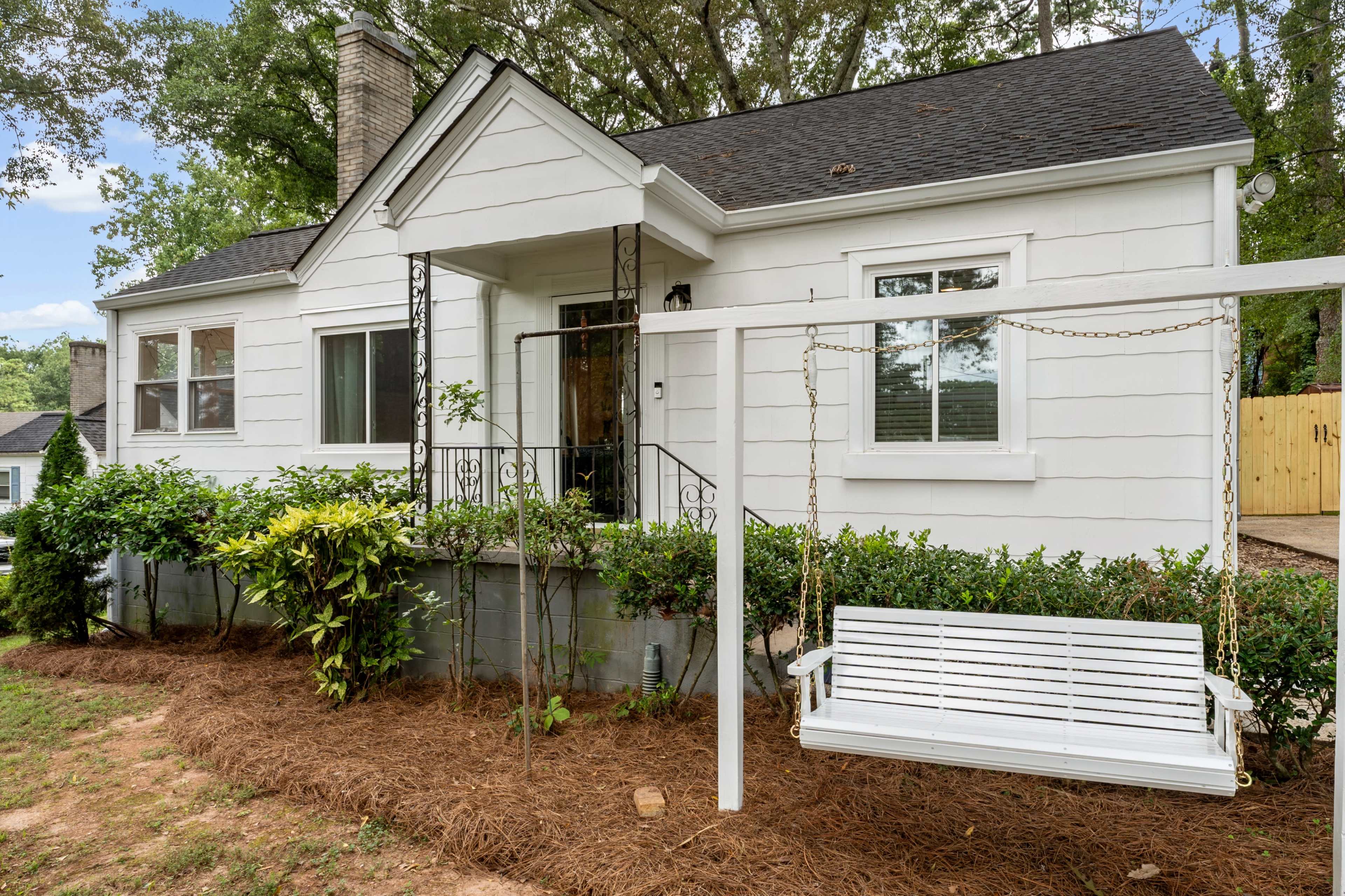 A white, single-story house with a front porch and a swing seat is surrounded by greenery and mulch.