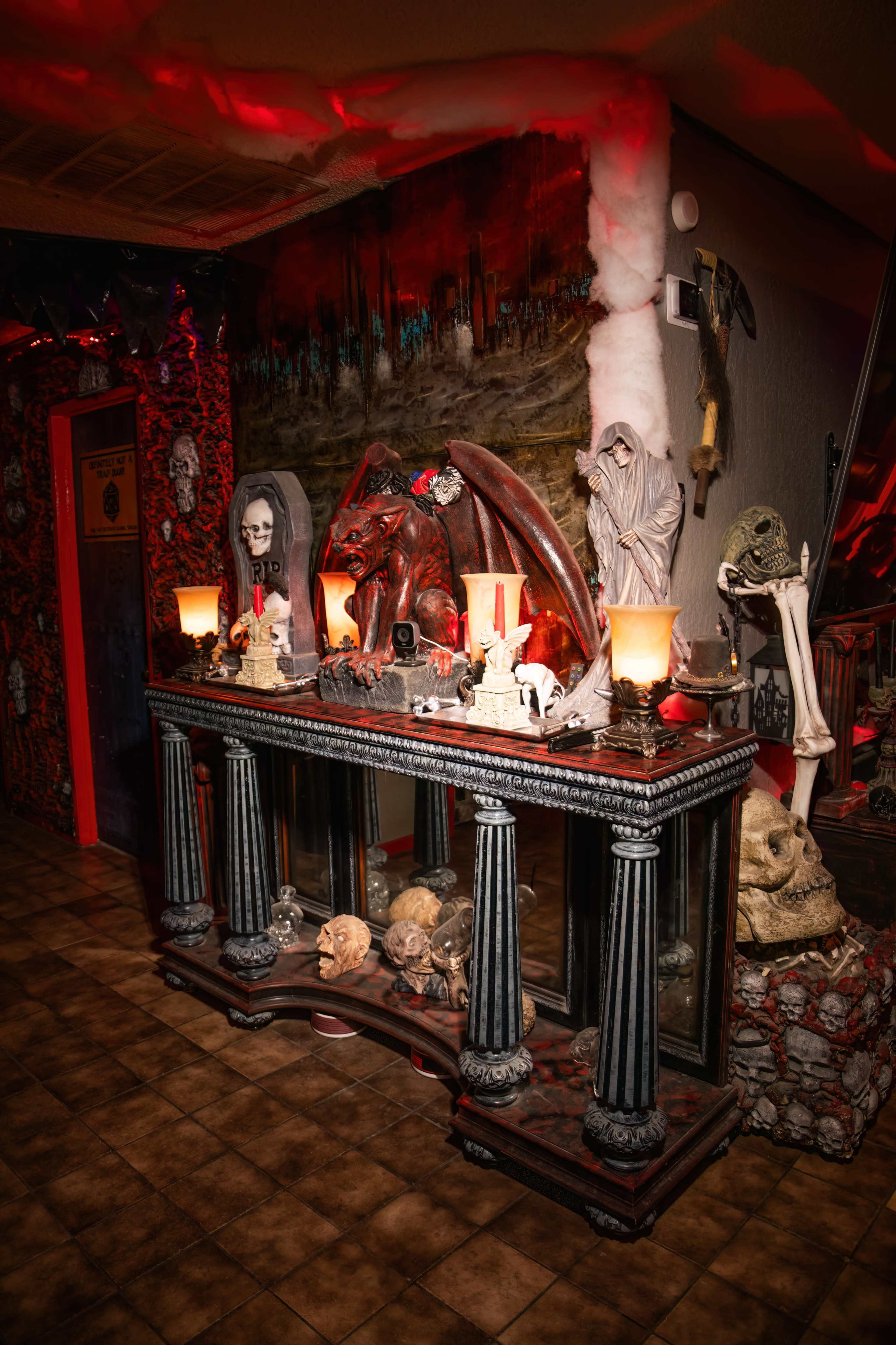 The scene depicts a dimly lit display area adorned with various horror-themed decorations, including a demonic figure, skulls, and eerie statues, along with flickering candles.