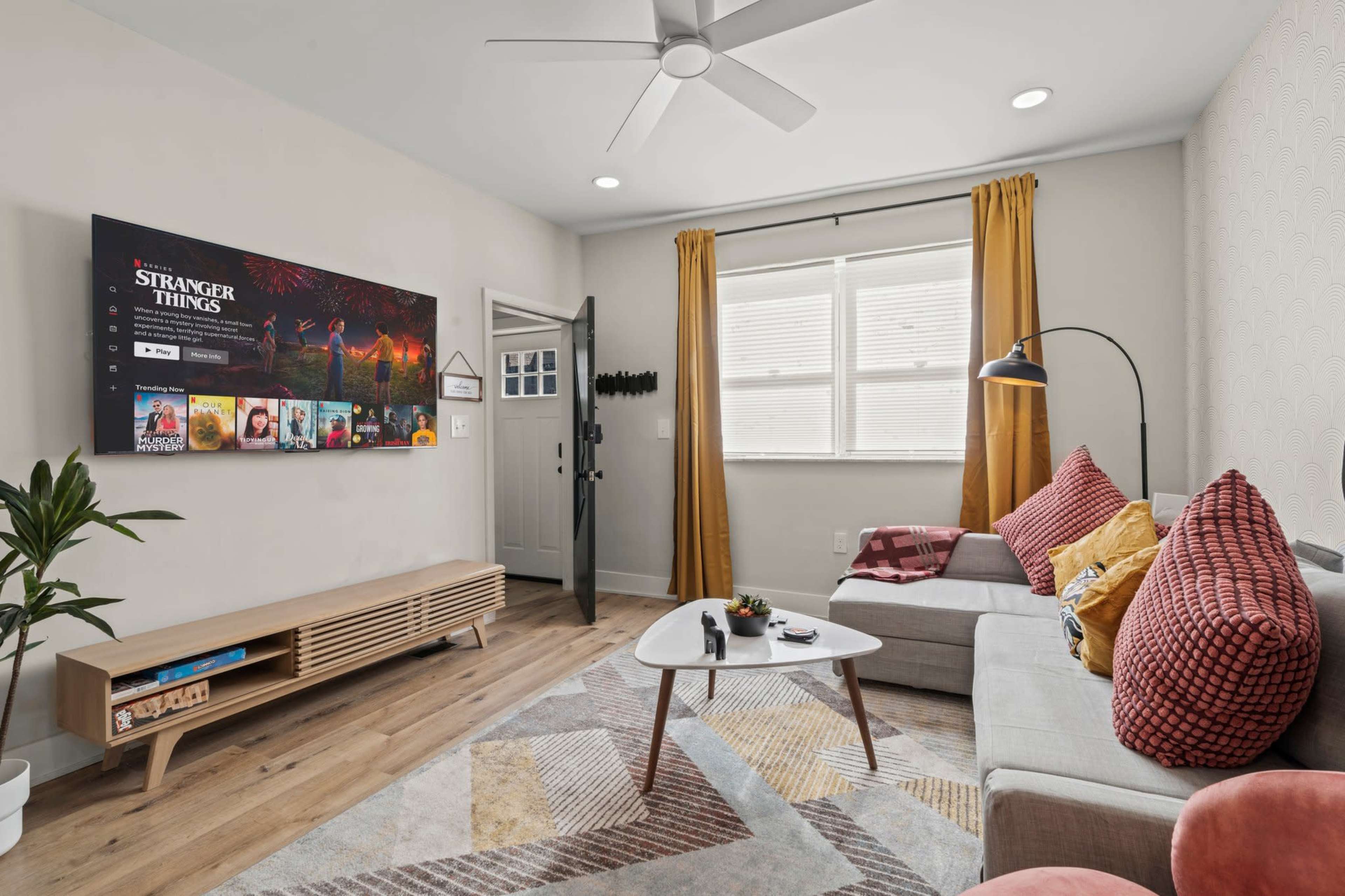 A modern living room features a large TV mounted on the wall, a gray sofa with pink cushions, a wooden coffee table, and an open door leading outside.