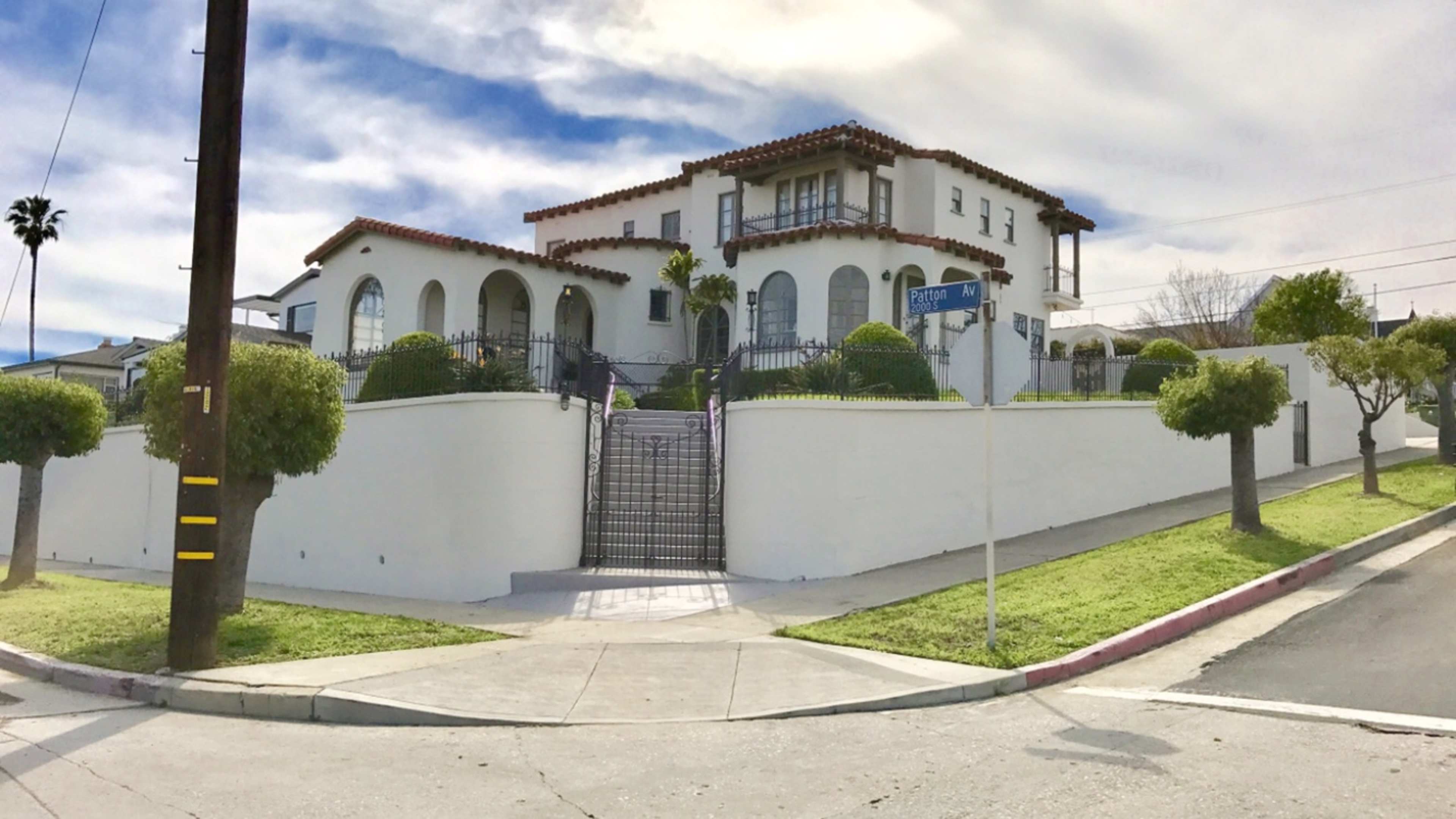 A large, two-story white house with a tiled roof sits on a landscaped corner lot, surrounded by a low white wall and ornamental trees.