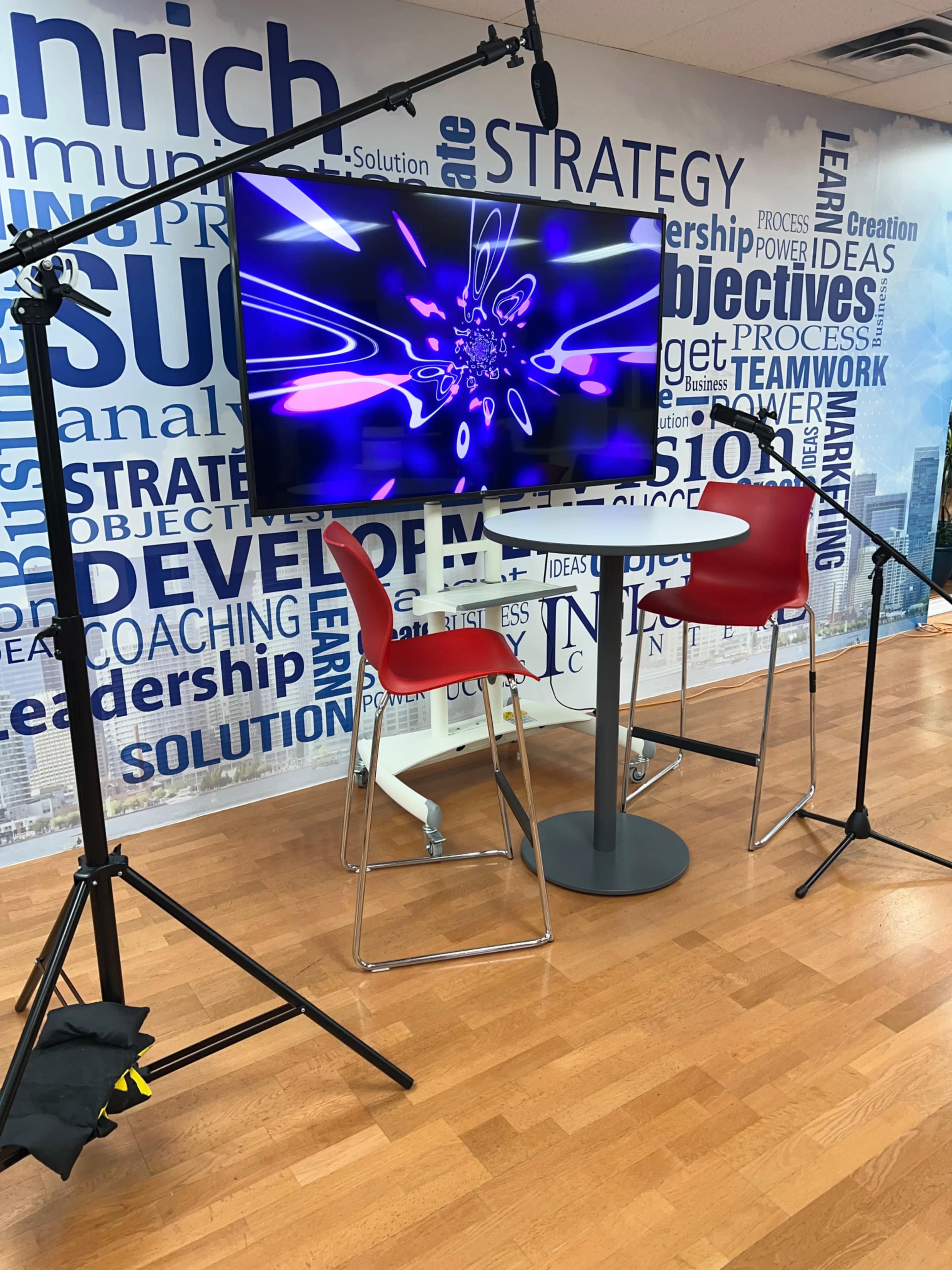 A sleek setup featuring a tall table and two red chairs in front of a large screen displaying abstract purple visuals, surrounded by a backdrop of business-related words.