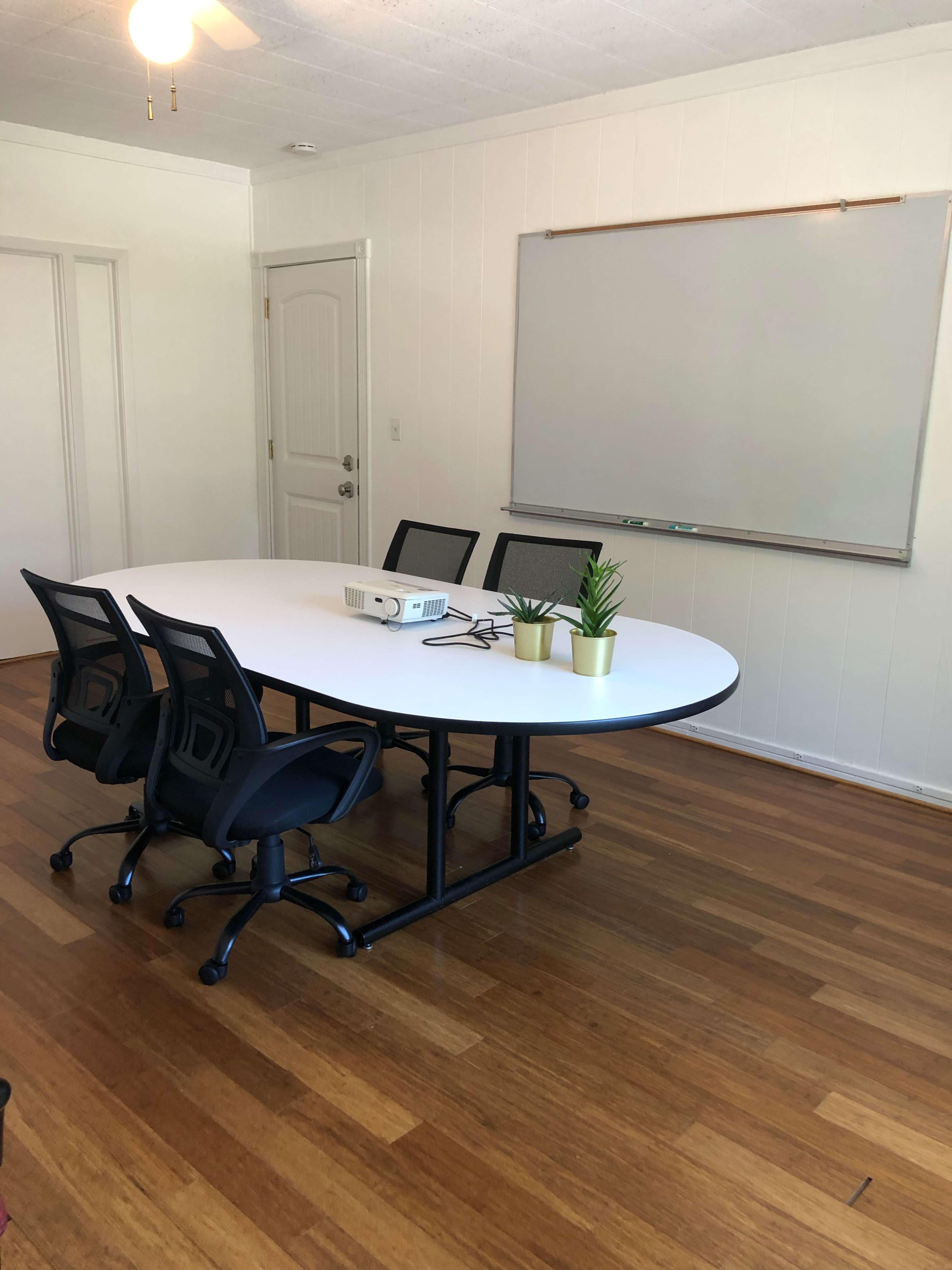 A conference room features a circular table surrounded by four black chairs, with a whiteboard and a projector in the background.