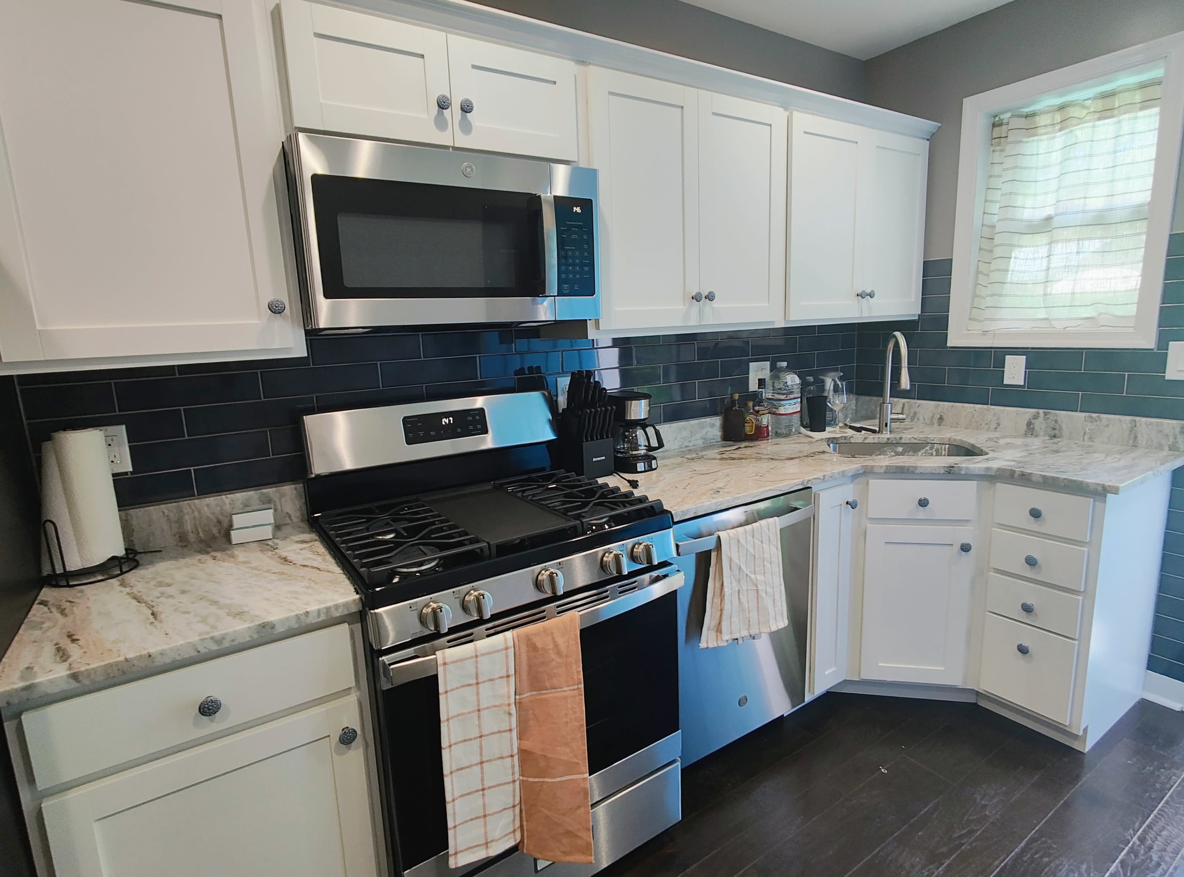 The image shows a modern kitchen featuring white cabinetry, a stainless steel refrigerator and microwave, a gas stove, and a granite countertop with a window above the sink.