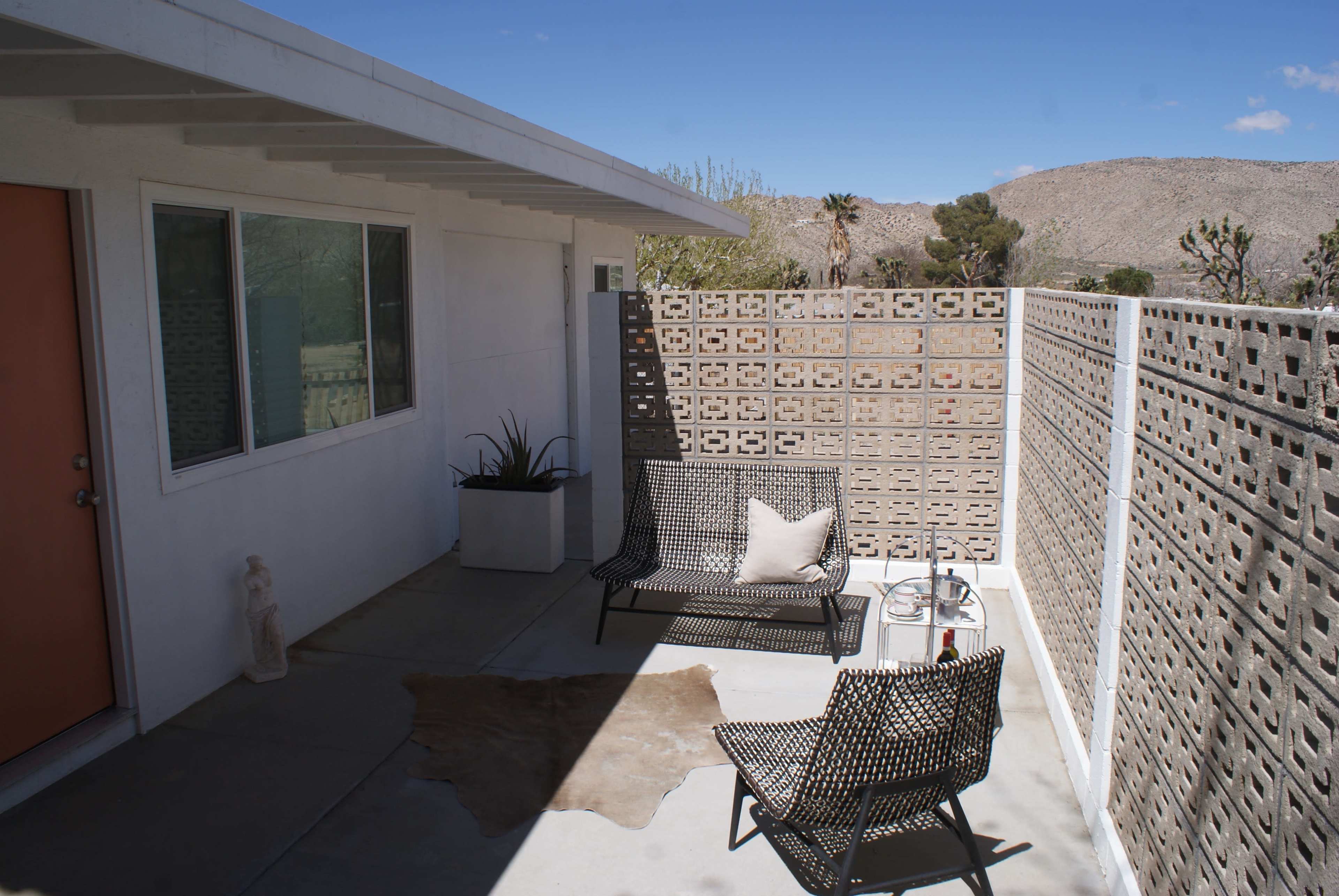A small outdoor patio area featuring two woven chairs, a small table, and a textured block wall, with a view of a desert landscape in the background.