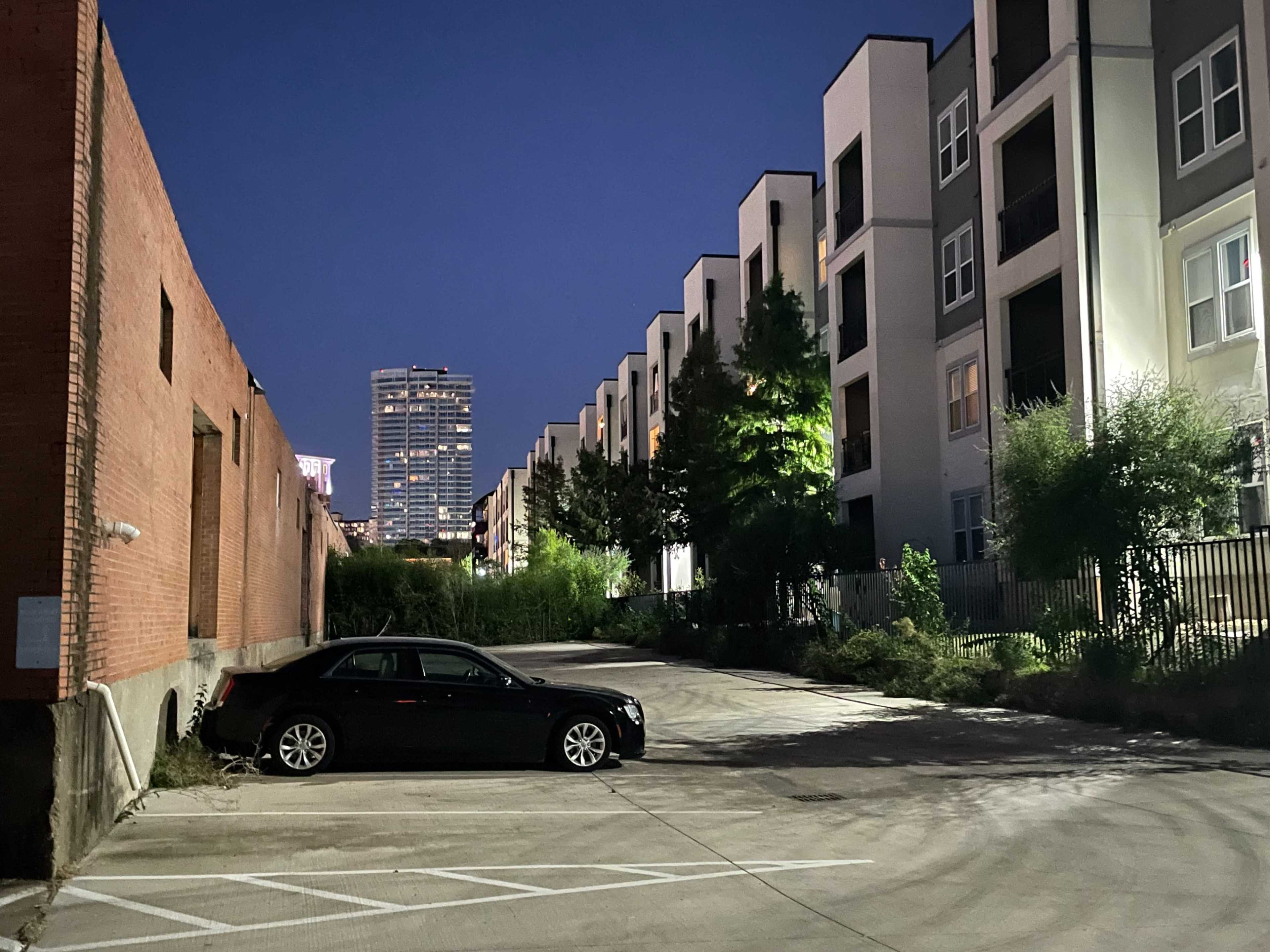 A black car is parked in an empty lot beside a row of modern apartment buildings at night, with a city skyline in the background.