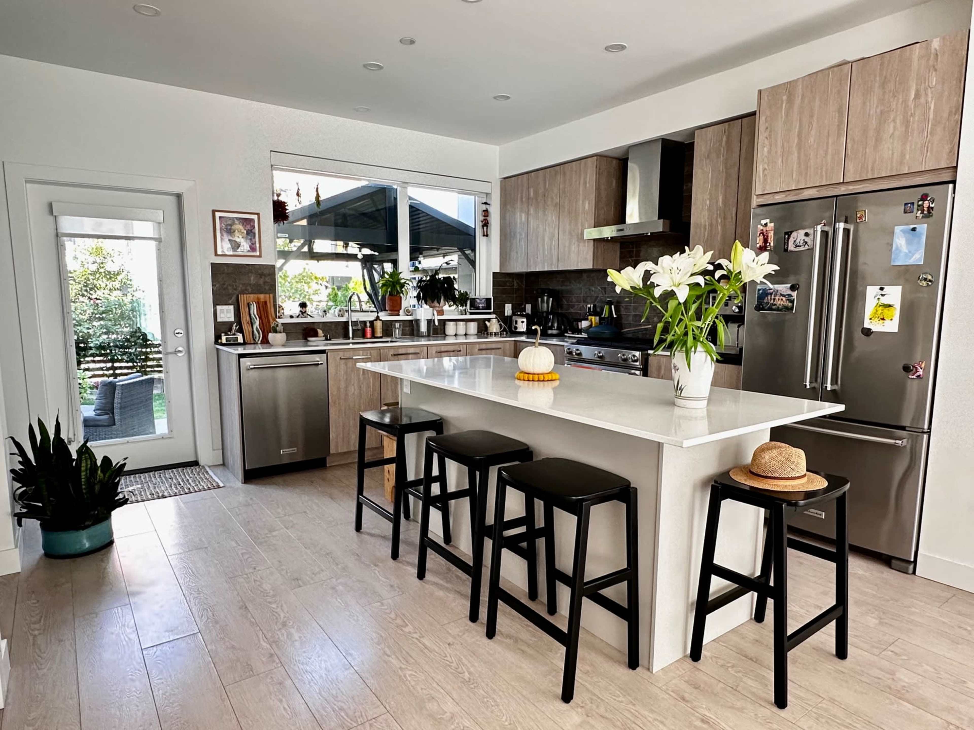 A modern kitchen features a large island with black stools, stainless steel appliances, and a view of a garden through the open door.