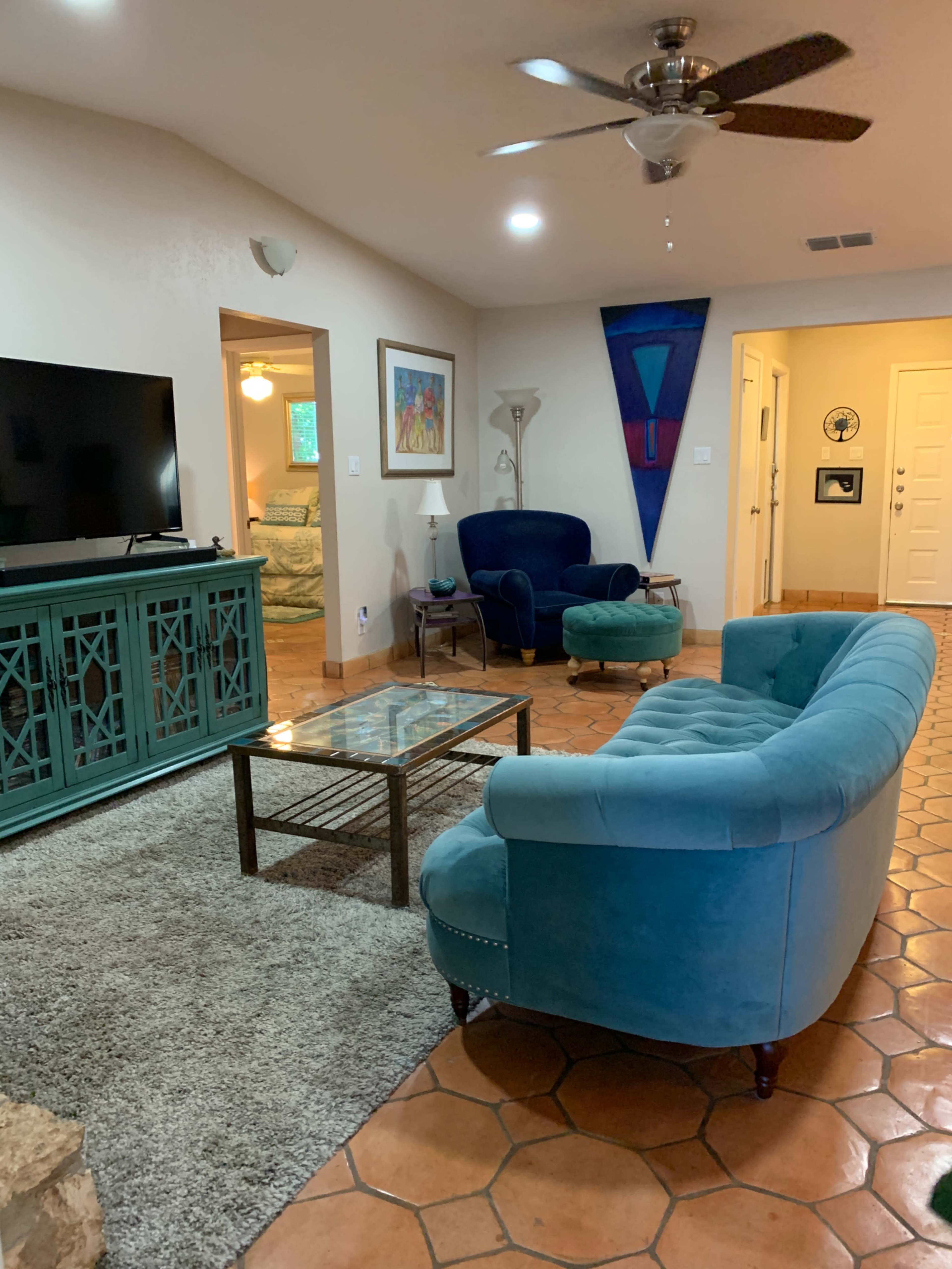 The living room features a teal sofa and armchair, a glass coffee table, a television, and a doorway leading to another room with a bed visible.