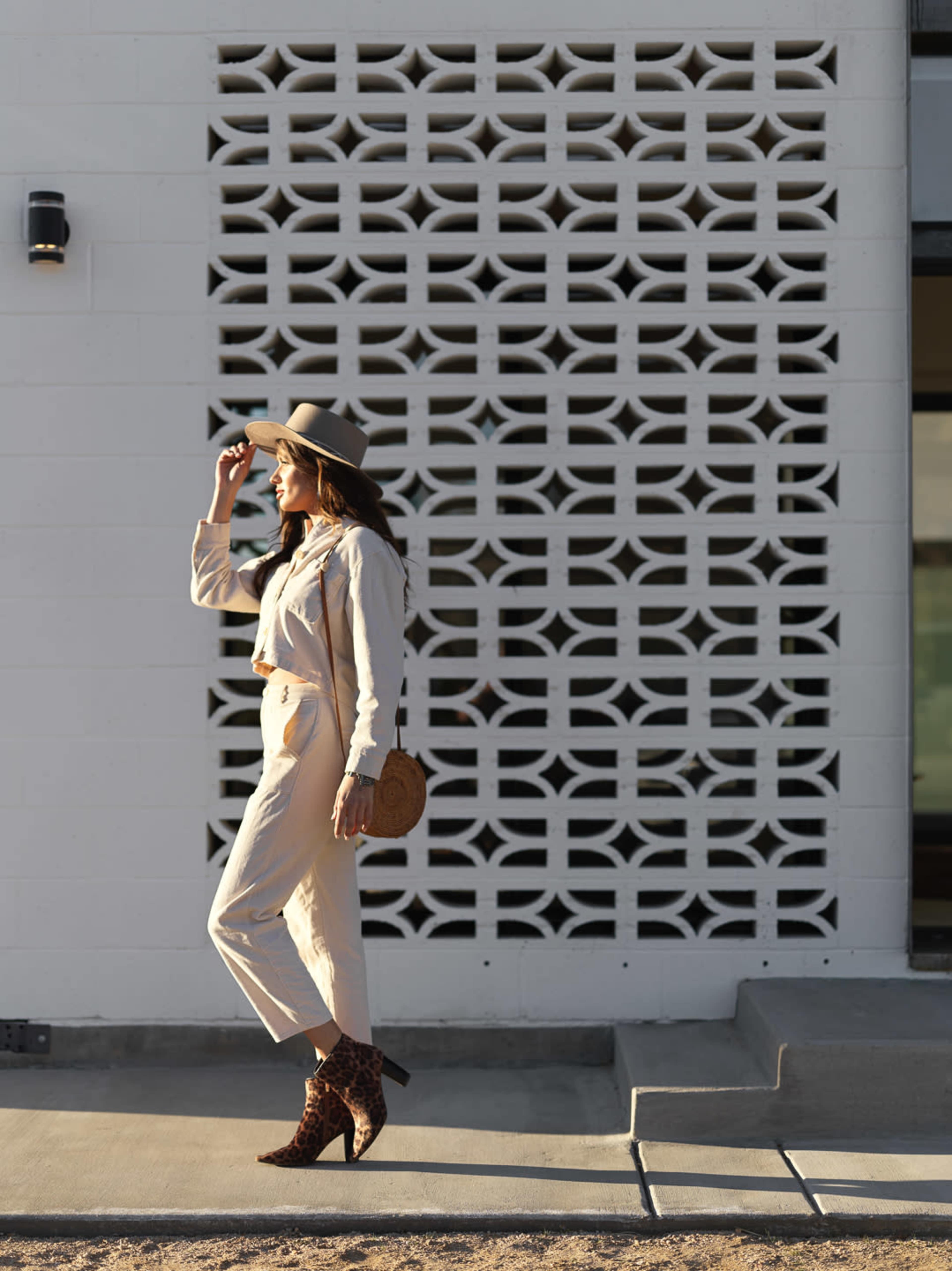 A woman in a light-colored outfit and hat walks beside a textured white wall with a geometric design.