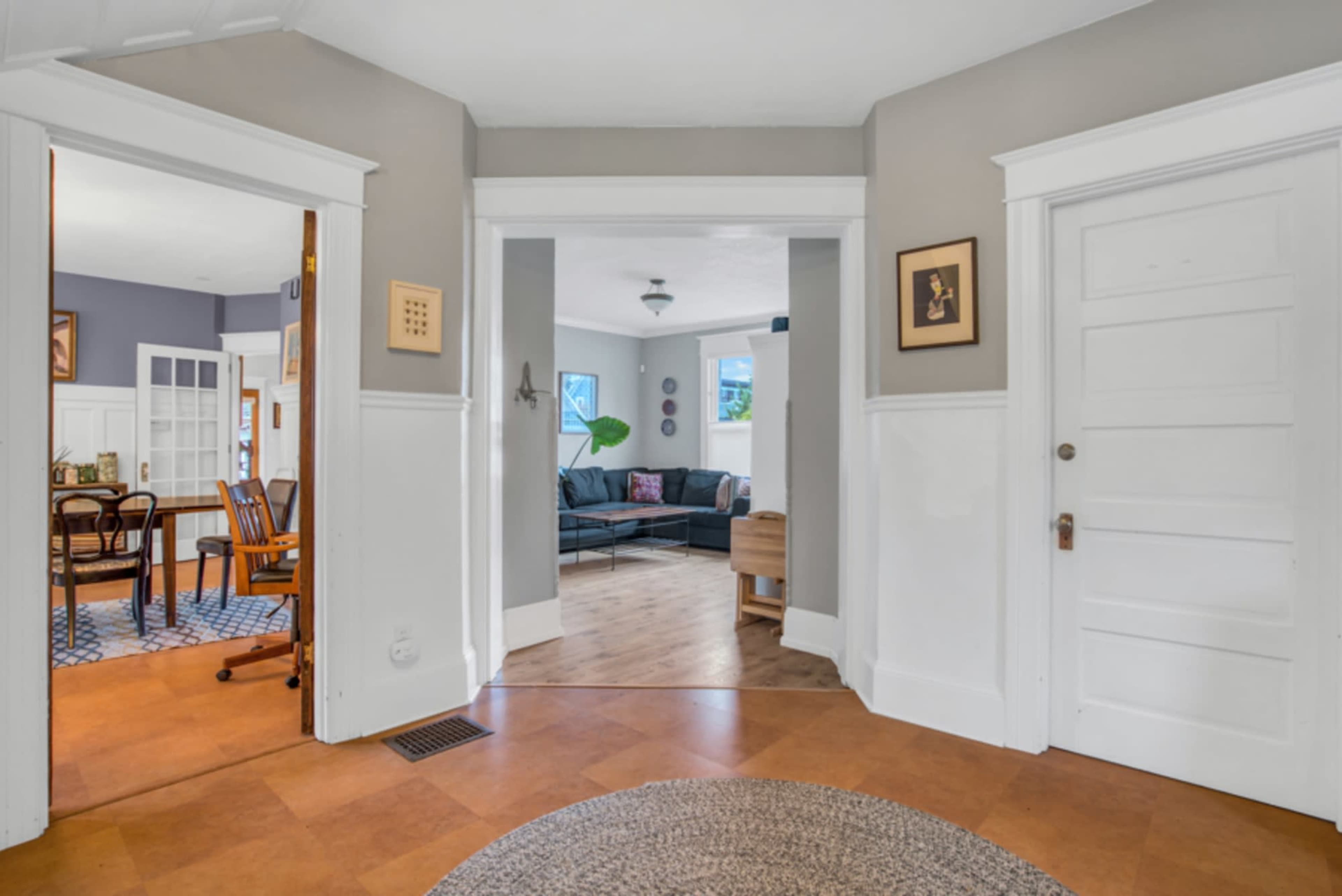 The image shows a well-lit hallway with a view into two adjacent rooms: a dining area on the left and a living room on the right, featuring large windows and wooden flooring.