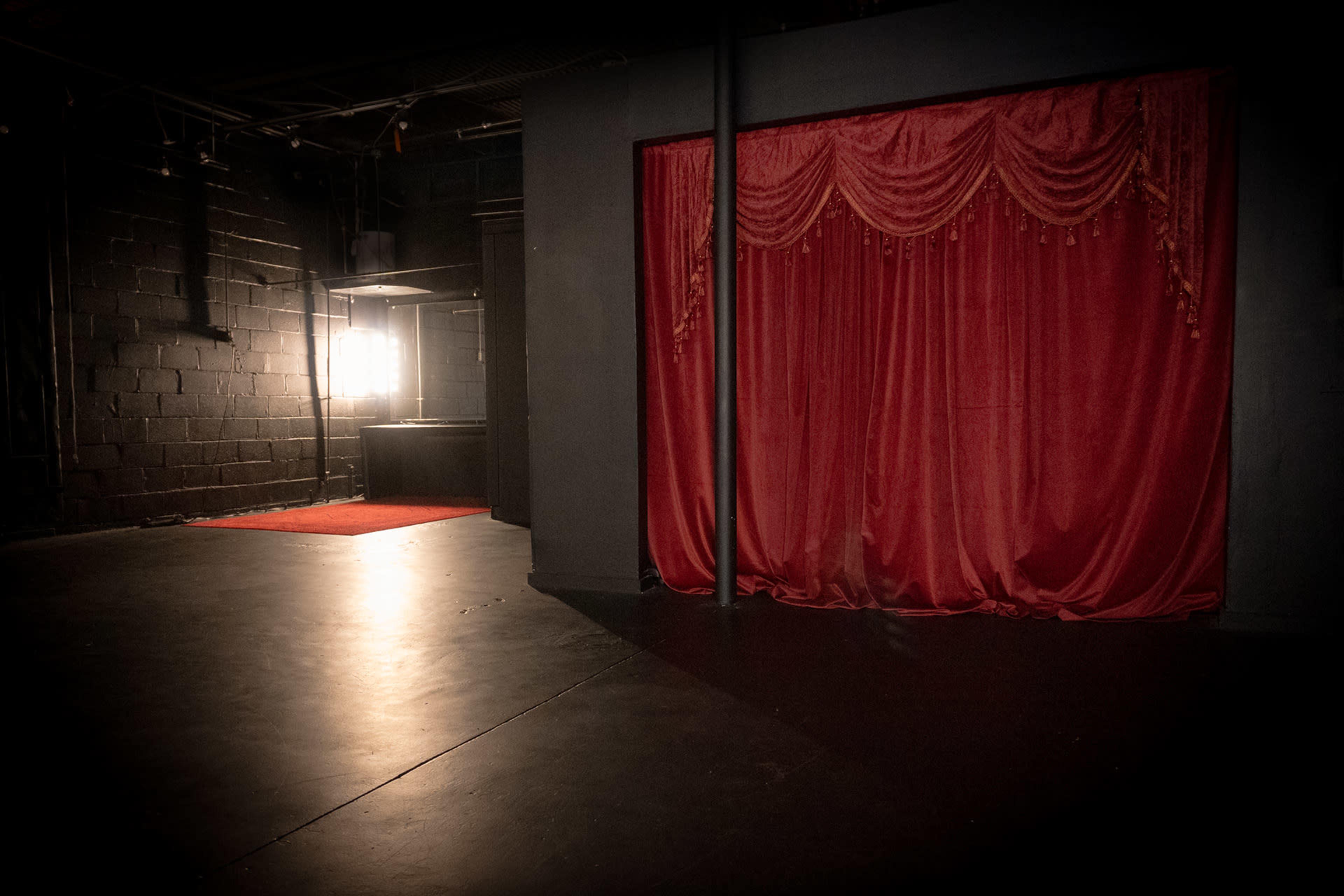 The image shows a dimly lit theater space featuring a red curtain on a stage, with a spotlight and a small vanity area visible in the background.
