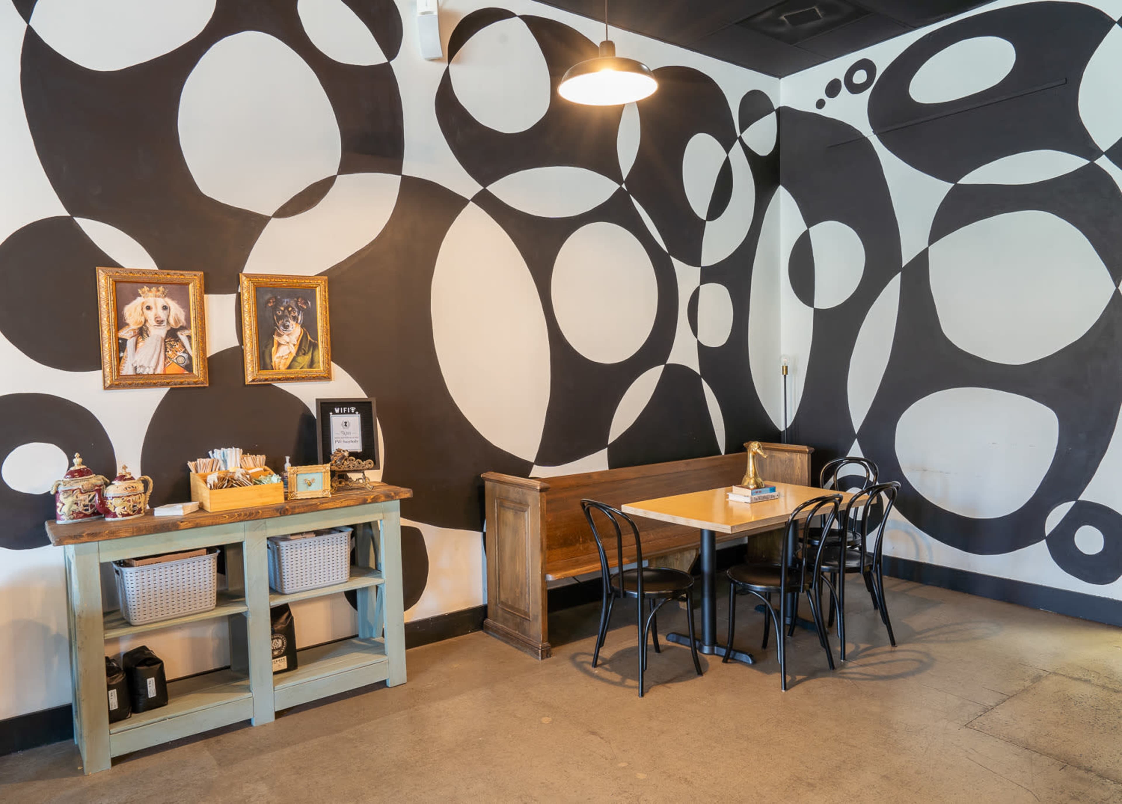 A café interior with a wooden table and chairs against a wall adorned with large black and white circular patterns, along with framed artwork and a service counter.