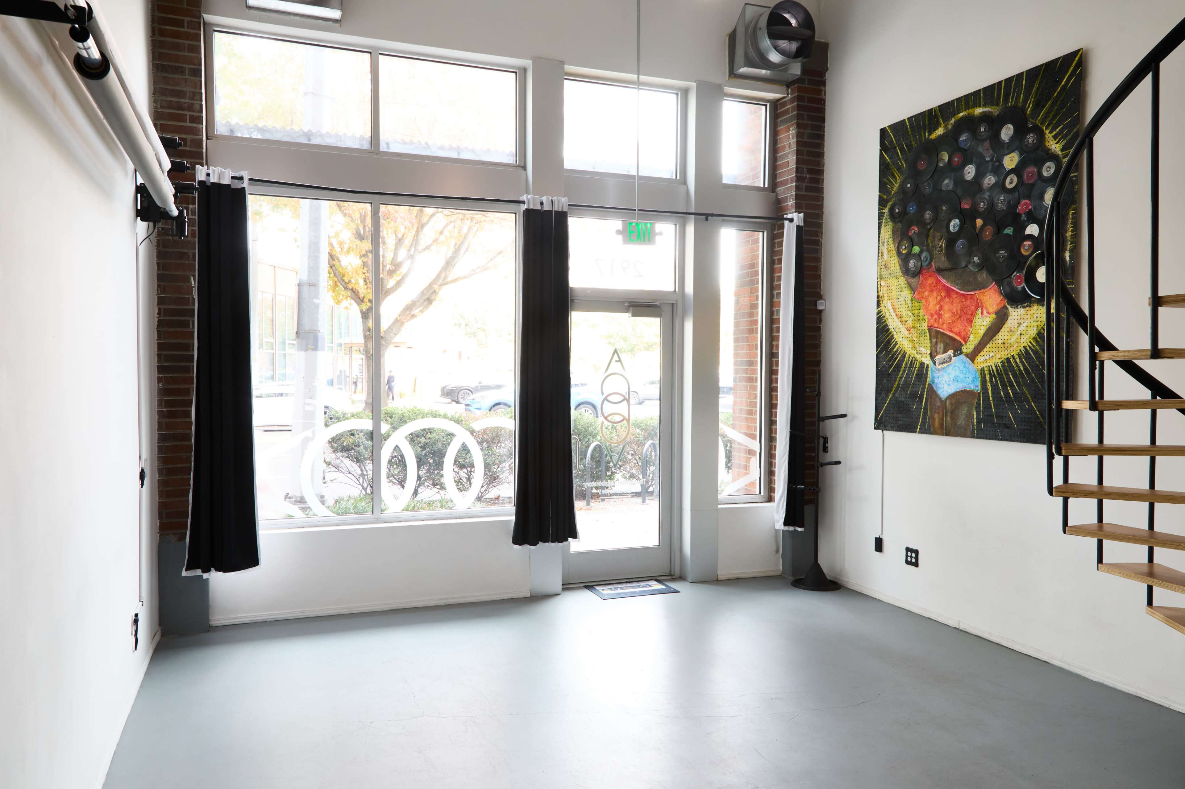 The interior of a bright studio features large windows with black curtains, a spiral staircase, and a colorful mural on the wall.