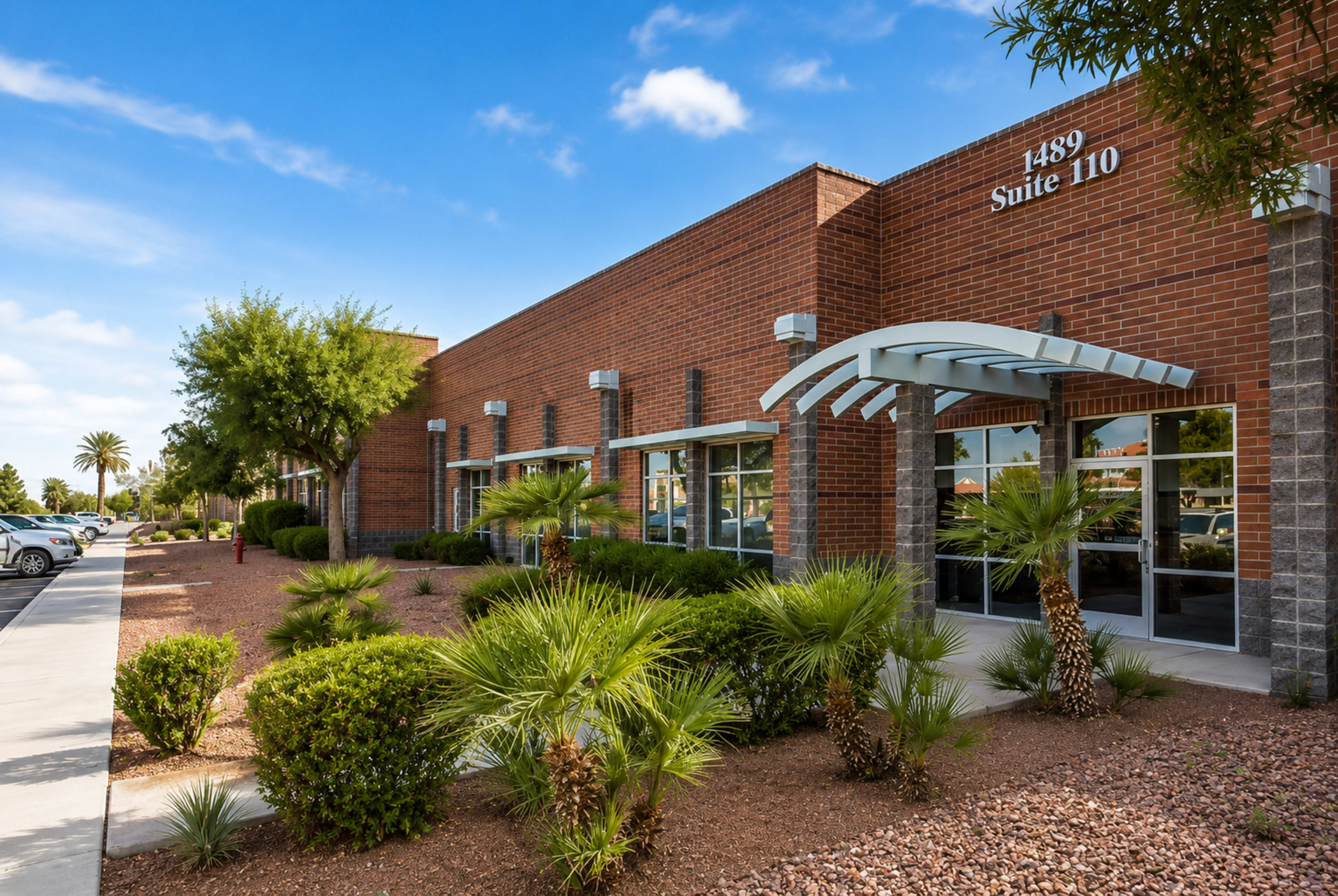 The image shows a modern brick building with large windows and landscaped shrubs in front, located in a pleasant outdoor setting.