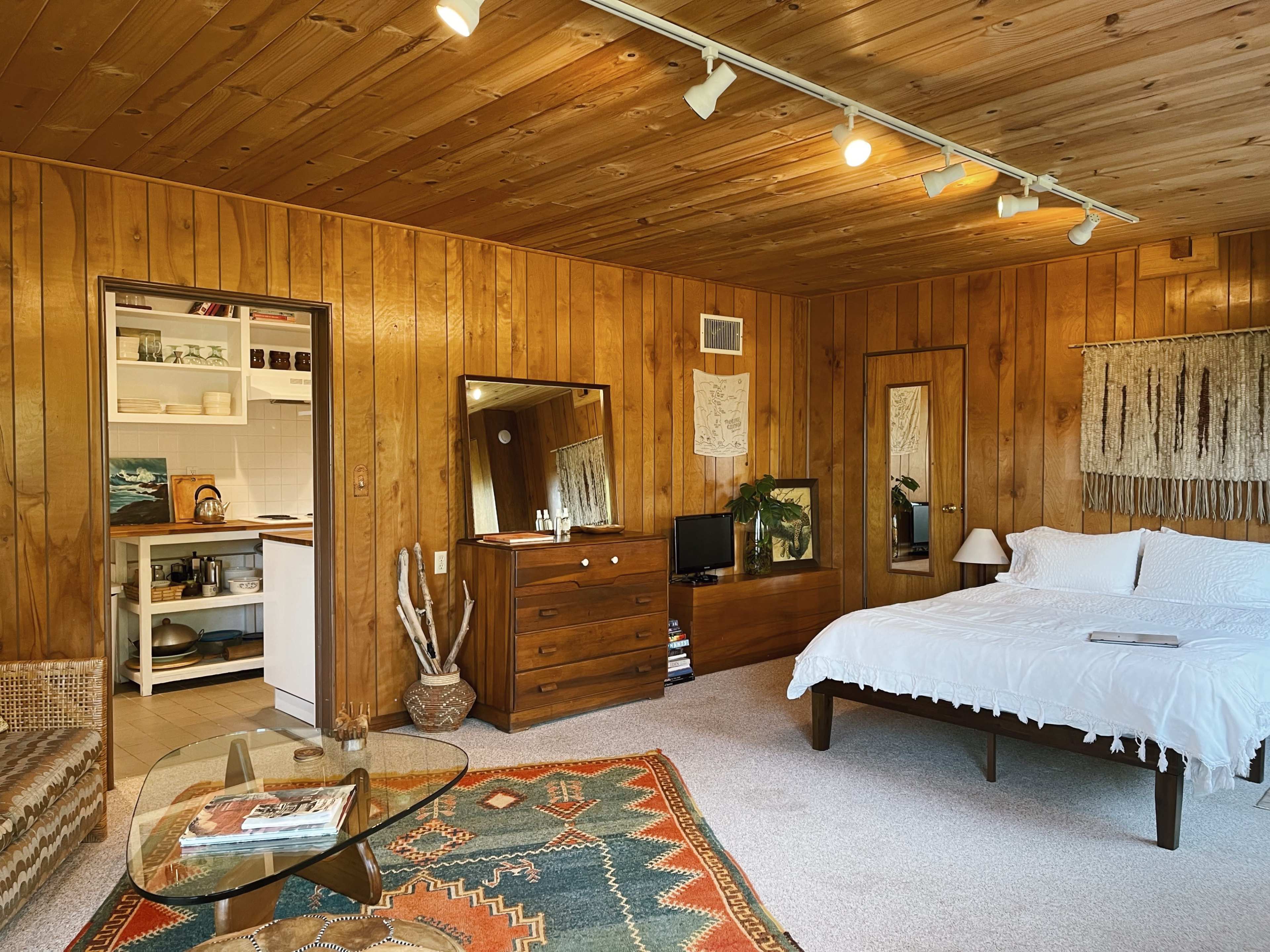 A wooden-paneled bedroom with a bed, a dresser, two mirrors, and a small kitchenette visible through an open doorway.