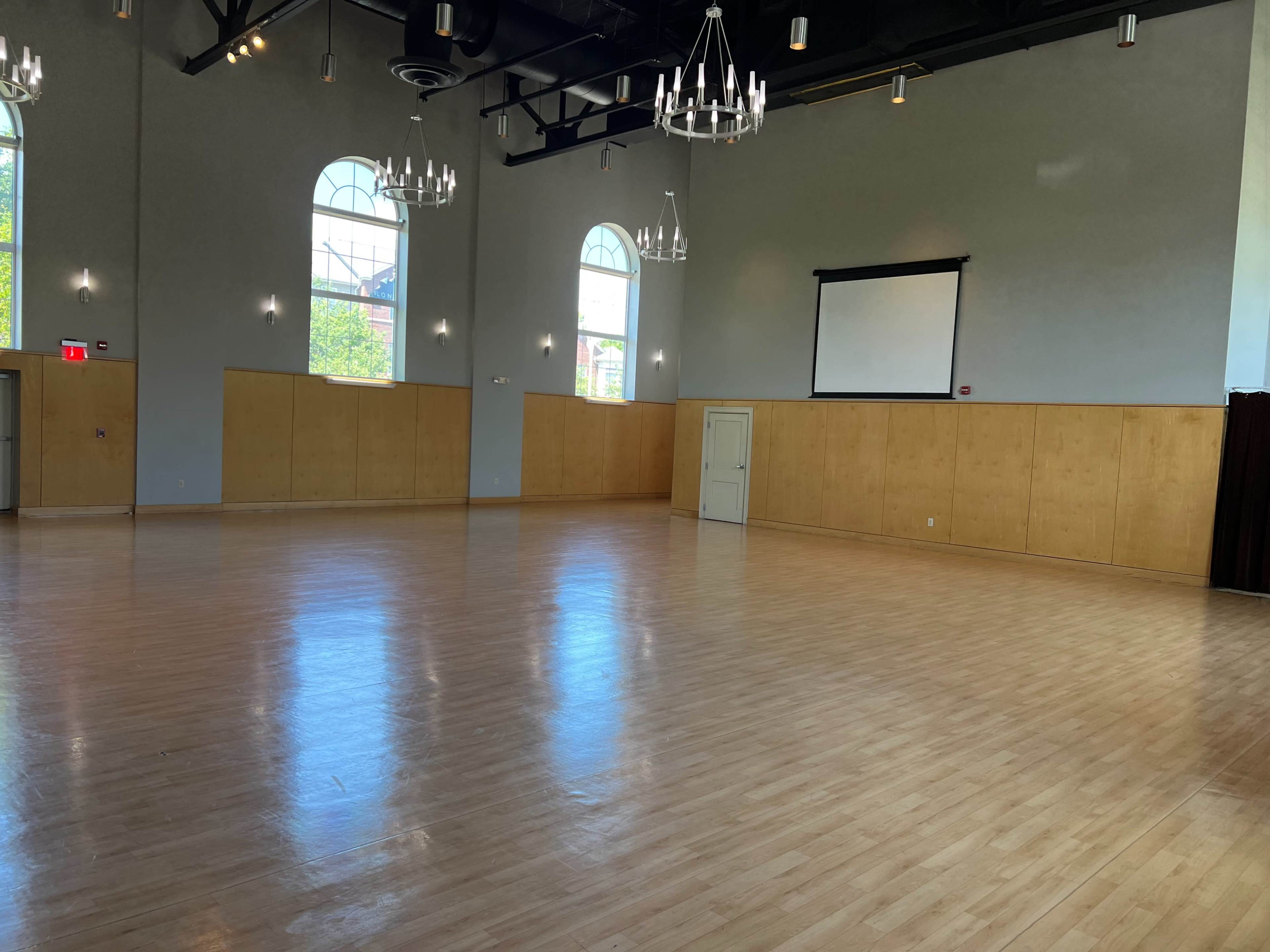 A spacious empty hall with wooden flooring, tall arched windows, and chandeliers hanging from the ceiling.