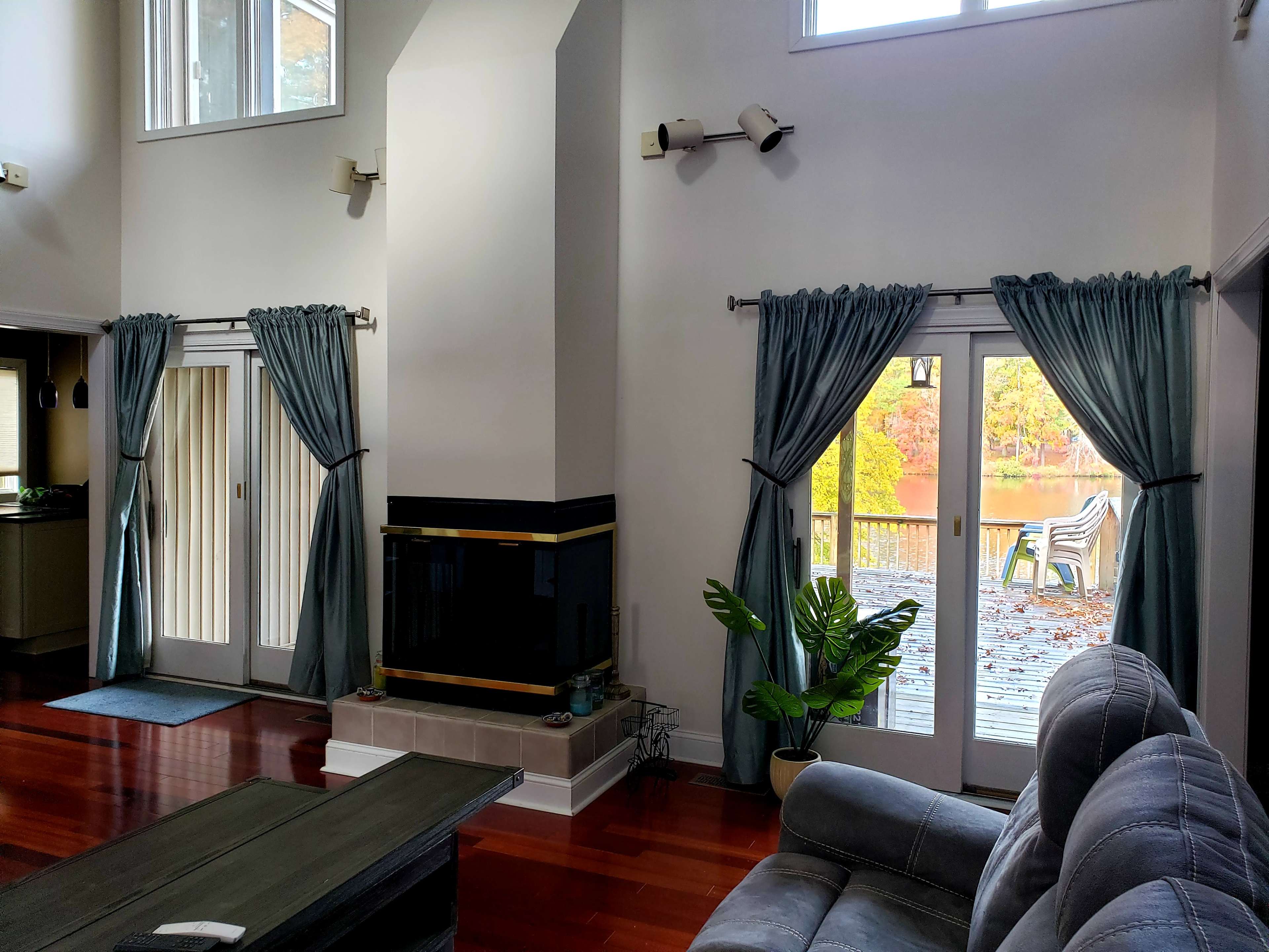 The interior of a living room features large windows with drapes, a fireplace, and a view of a deck with outdoor seating by a body of water.
